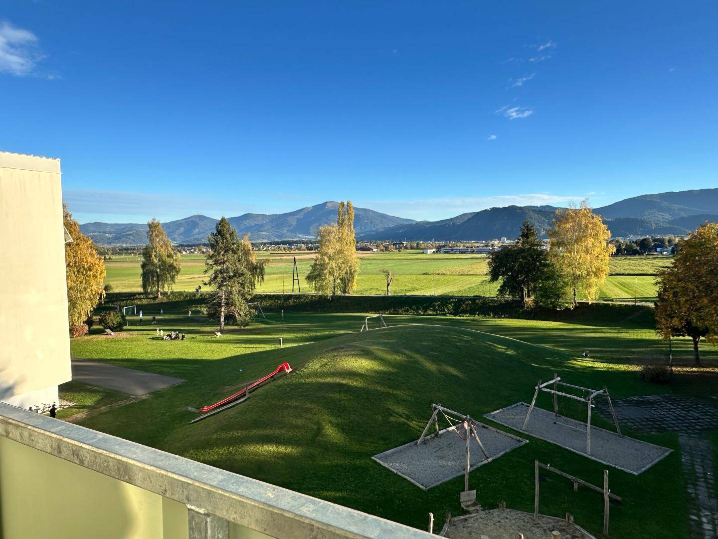 Balkon mit Blick auf einen Spielplatz und weite grüne Wiesen mit Bergen im Hintergrund.