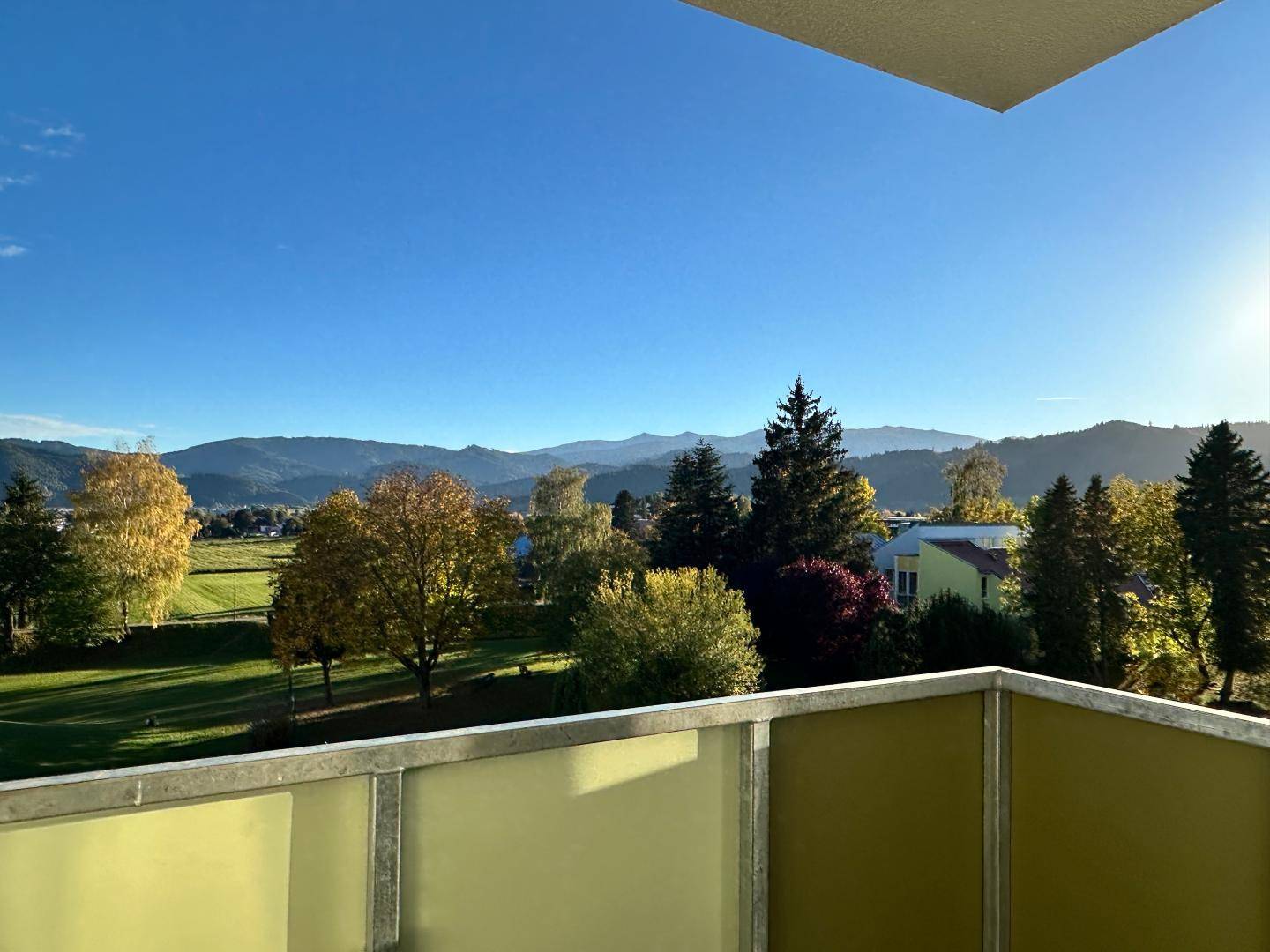 Balkon mit weitem Blick über grüne Felder und Berge unter blauem Himmel.