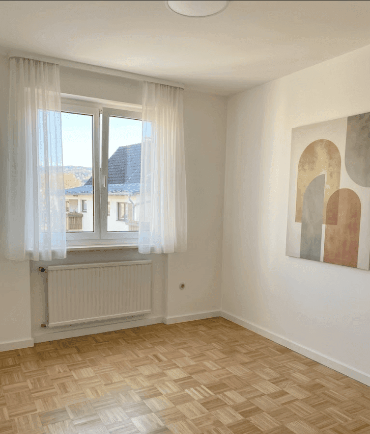 Heller Raum mit Parkettboden, Fenster und einem modernen Kunstwerk an der Wand.