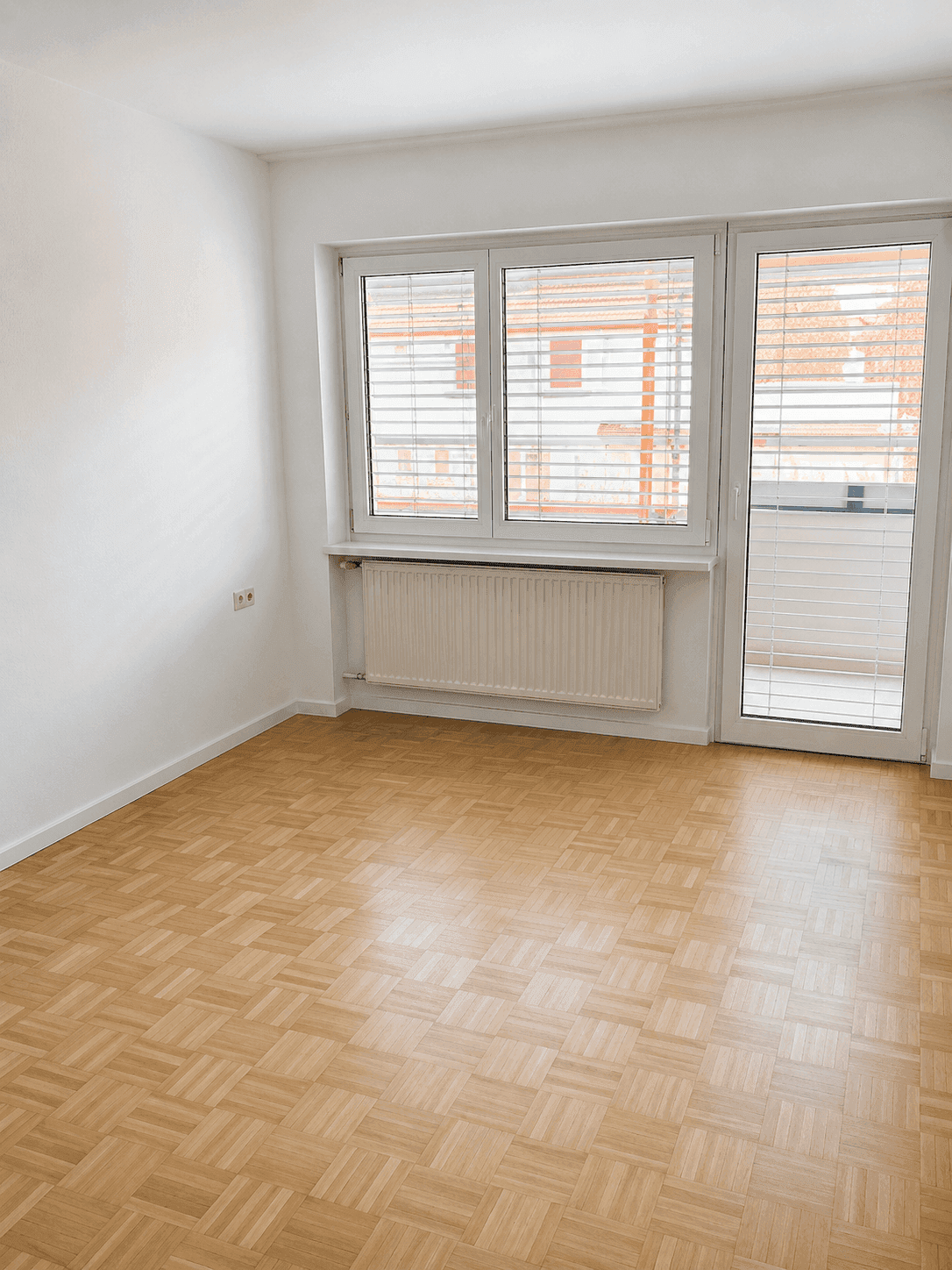 Heller Raum mit Parkettboden, großem Fenster und Zugang zu einem Balkon.