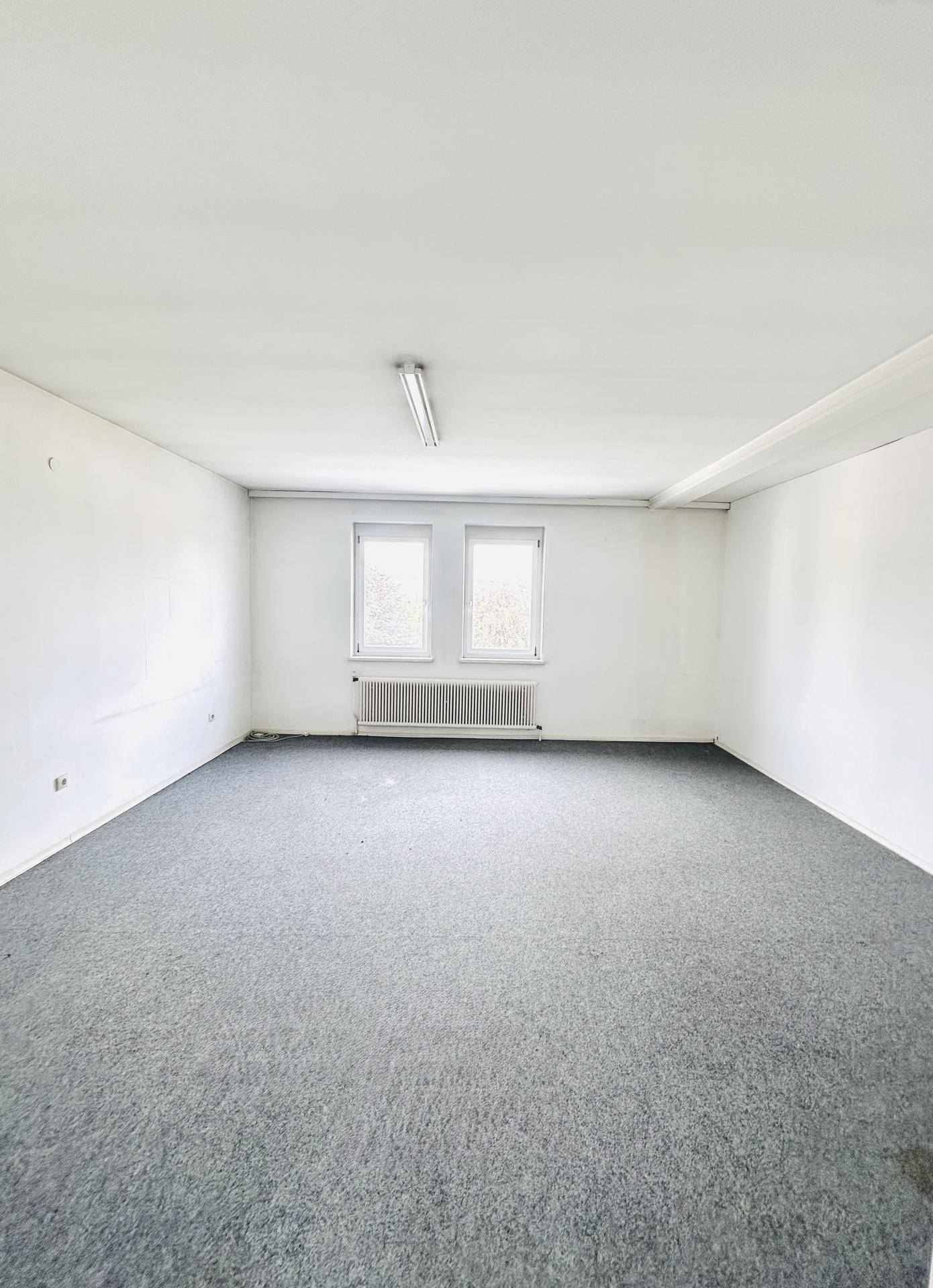 Leerer Büroraum mit grauem Teppichboden, weißen Wänden und zwei Fenstern, die Tageslicht spenden.