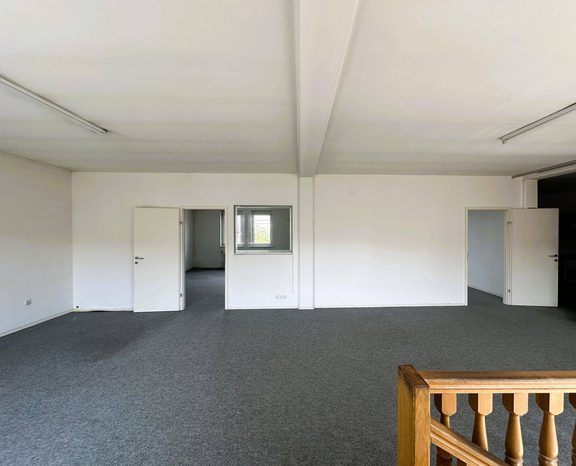 Obergeschoss-Büroraum mit grauem Teppichboden, weißen Wänden und Zugang zu weiteren Räumen.