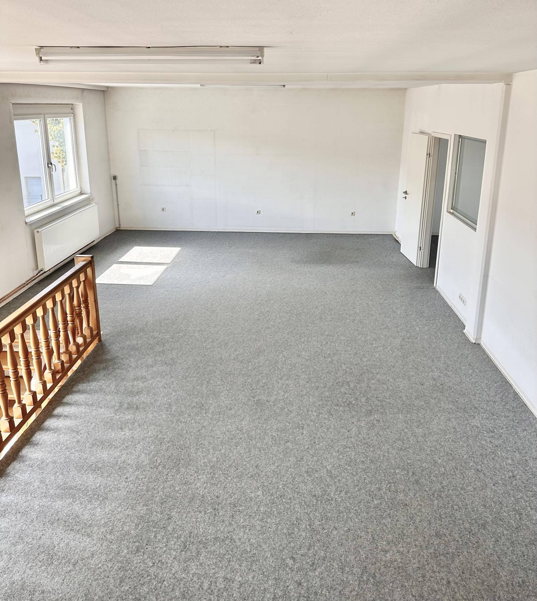 Heller Büroraum im Obergeschoss mit grauem Teppichboden, Fenster und Holzgeländer.