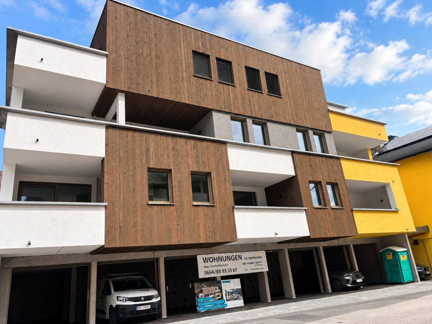 Modernes Mehrfamilienhaus mit Holzfassade und gelben Akzenten sowie Tiefgaragenzufahrt.
