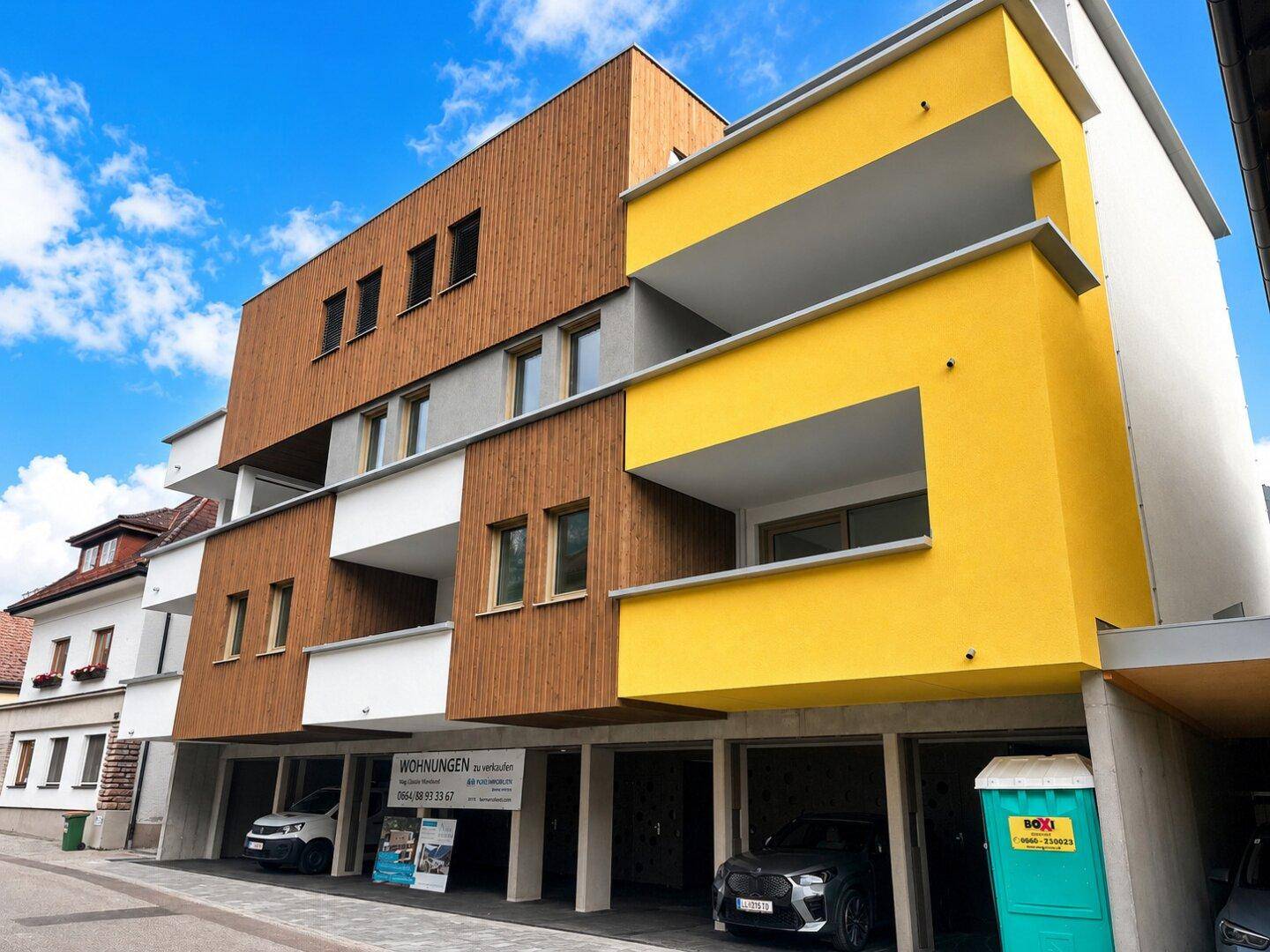 Modernes Wohnhaus mit markanter Holzfassade, gelben Balkonen und offener Tiefgarage.