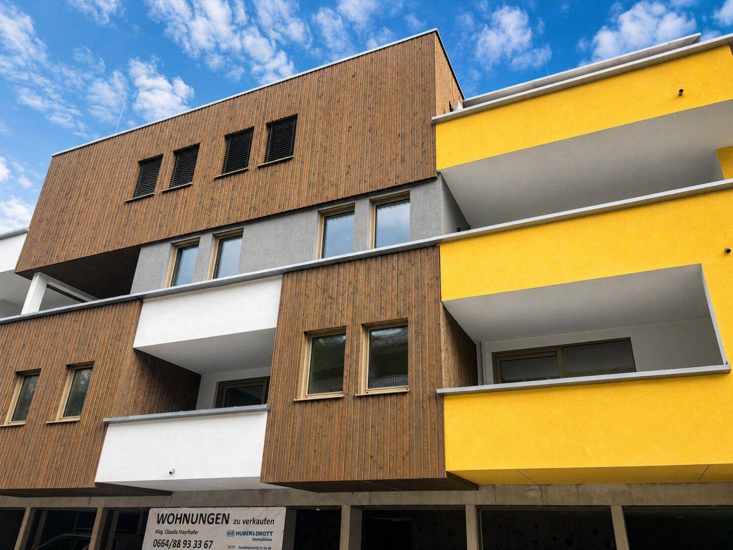 Fassade eines Neubaus mit Holzverkleidung, gelben Akzenten und modernen Balkonen.
