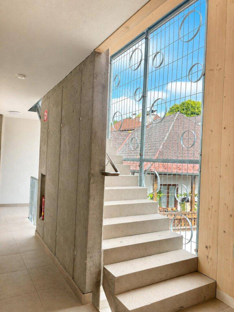 Heller Treppenaufgang mit Betonwand und großen Fenstern, die viel Licht hereinlassen.
