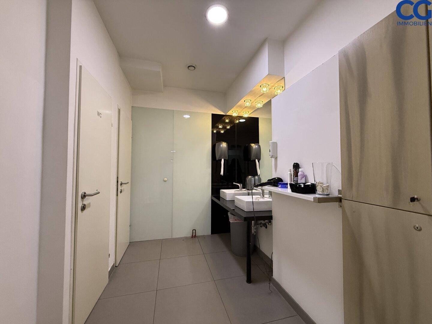 Waschraum mit zwei Waschbecken, großen Spiegeln und hellen Wänden, der zu den Toilettenkabinen führt.