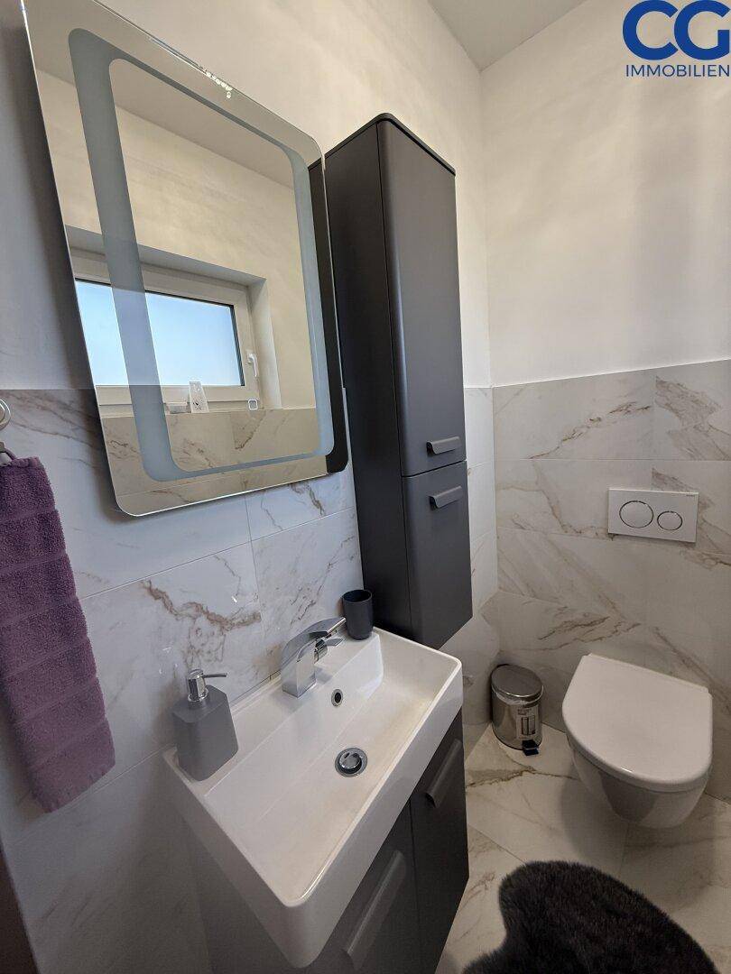 Gäste-WC mit modernen Fliesen, wandhängendem WC und beleuchtetem Spiegel.