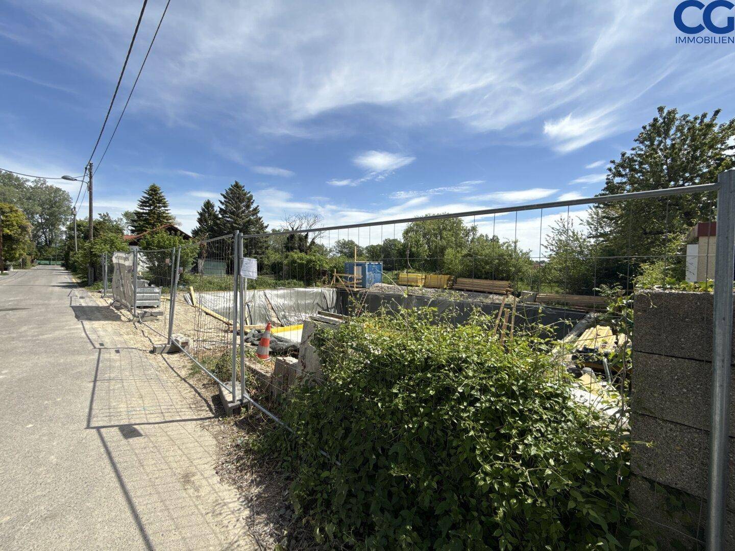 Blick von der Straße auf die umzäunte Baustelle mit umliegenden Bäumen und Büschen.