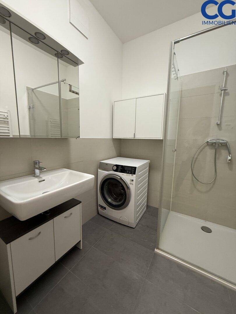 Funktionales Badezimmer mit ebenerdiger Dusche, Waschbecken, Spiegelschrank und Waschmaschine.