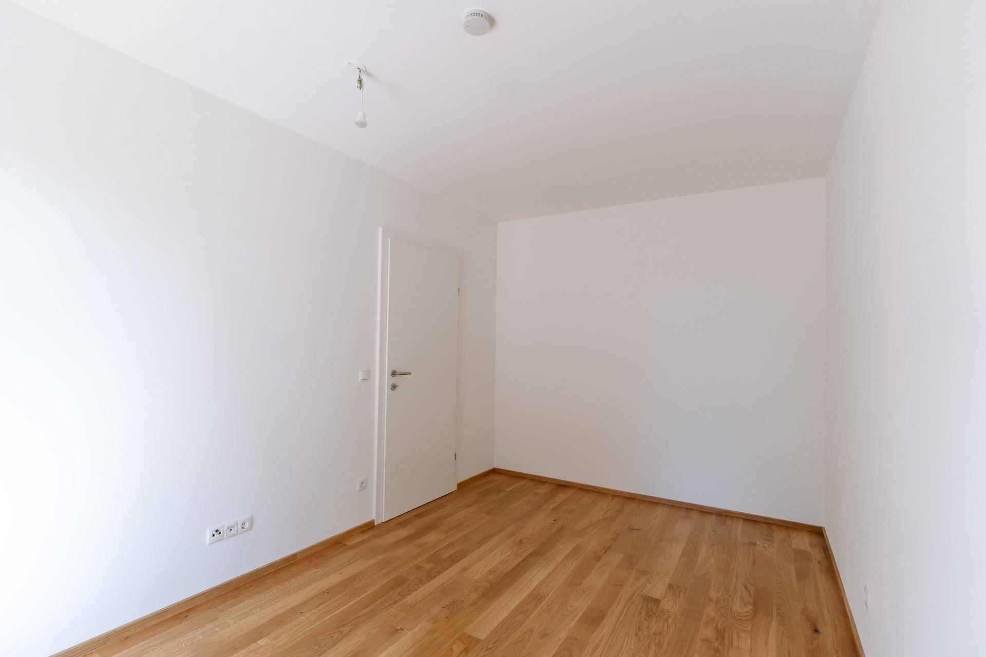 Helles, leeres Zimmer mit Parkettboden und weißer Tür, ideal als Schlaf- oder Arbeitszimmer.