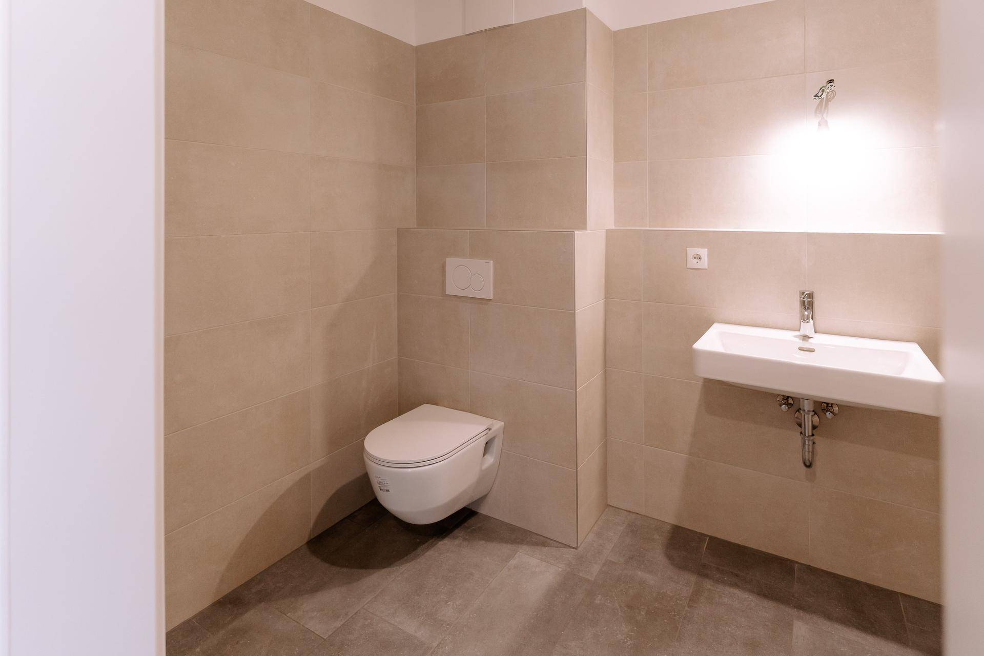 Separates WC mit wandhängender Toilette, Waschbecken und beigefarbenen Wandfliesen.