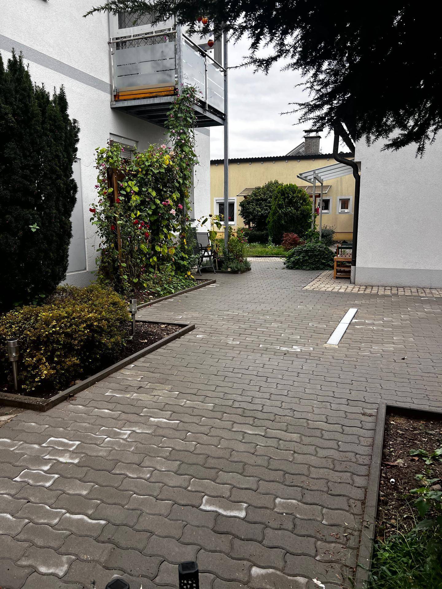 Gepflasterter Außenbereich mit Grünflächen und einem kleinen Balkon im Hintergrund.