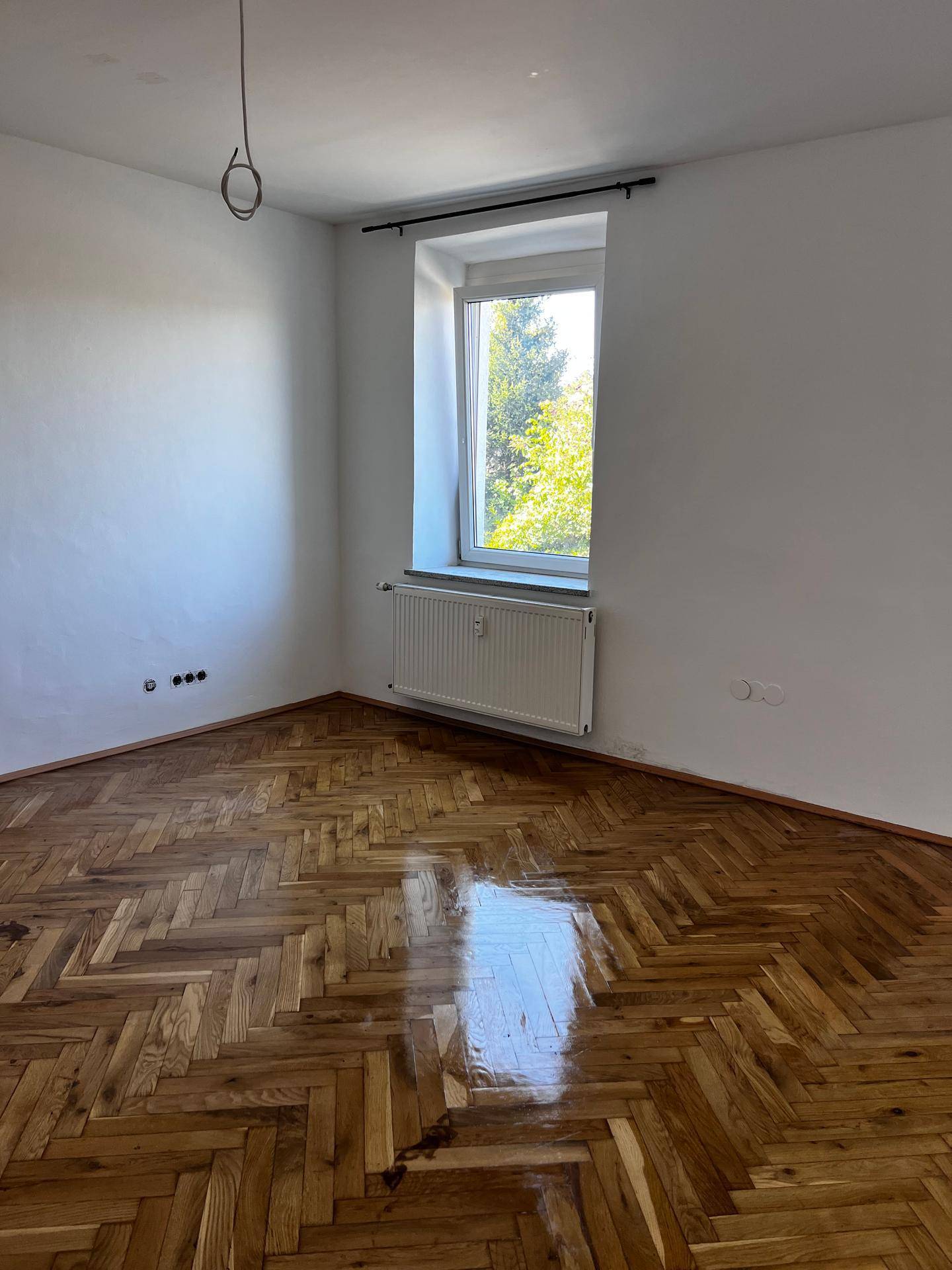 Lichtdurchflutetes Zimmer mit glänzendem Fischgrätparkett und Blick ins Grüne.