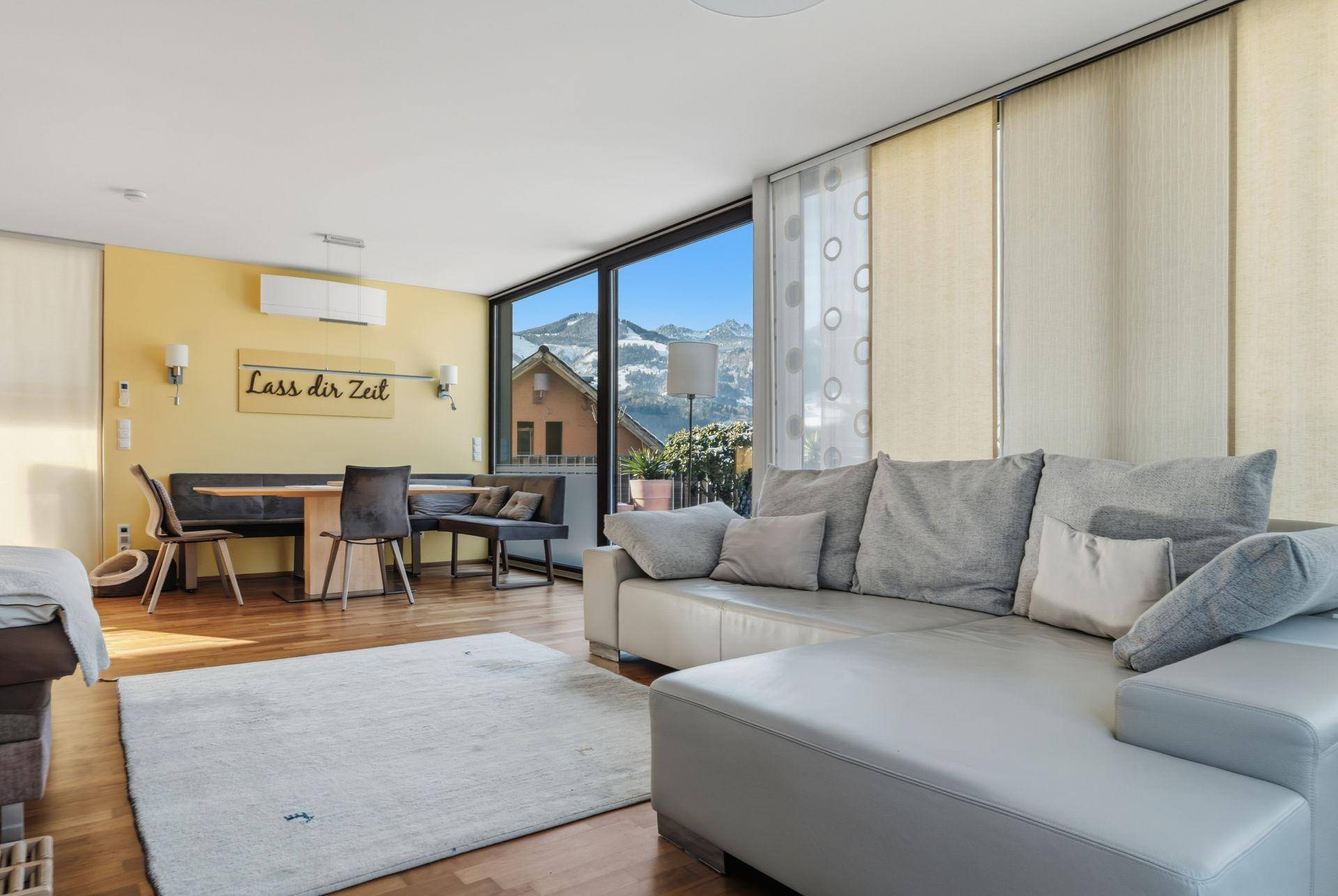 Helles Wohn-Esszimmer mit großem Sofa, Essbereich und Panoramafenstern mit Bergblick.