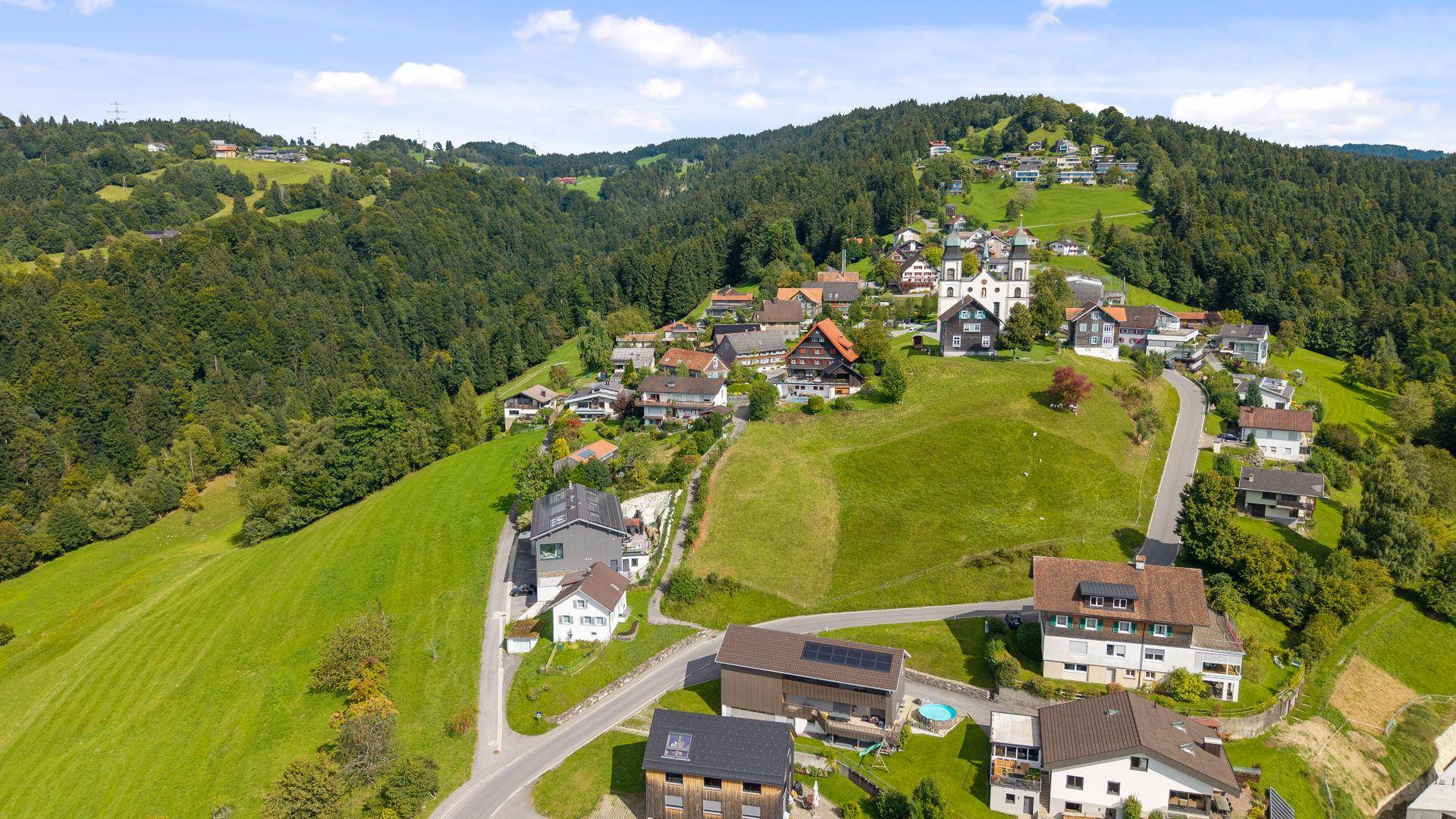 Luftaufnahme eines malerischen Dorfes in grüner Hügellandschaft mit Kirche und Wohnhäusern.