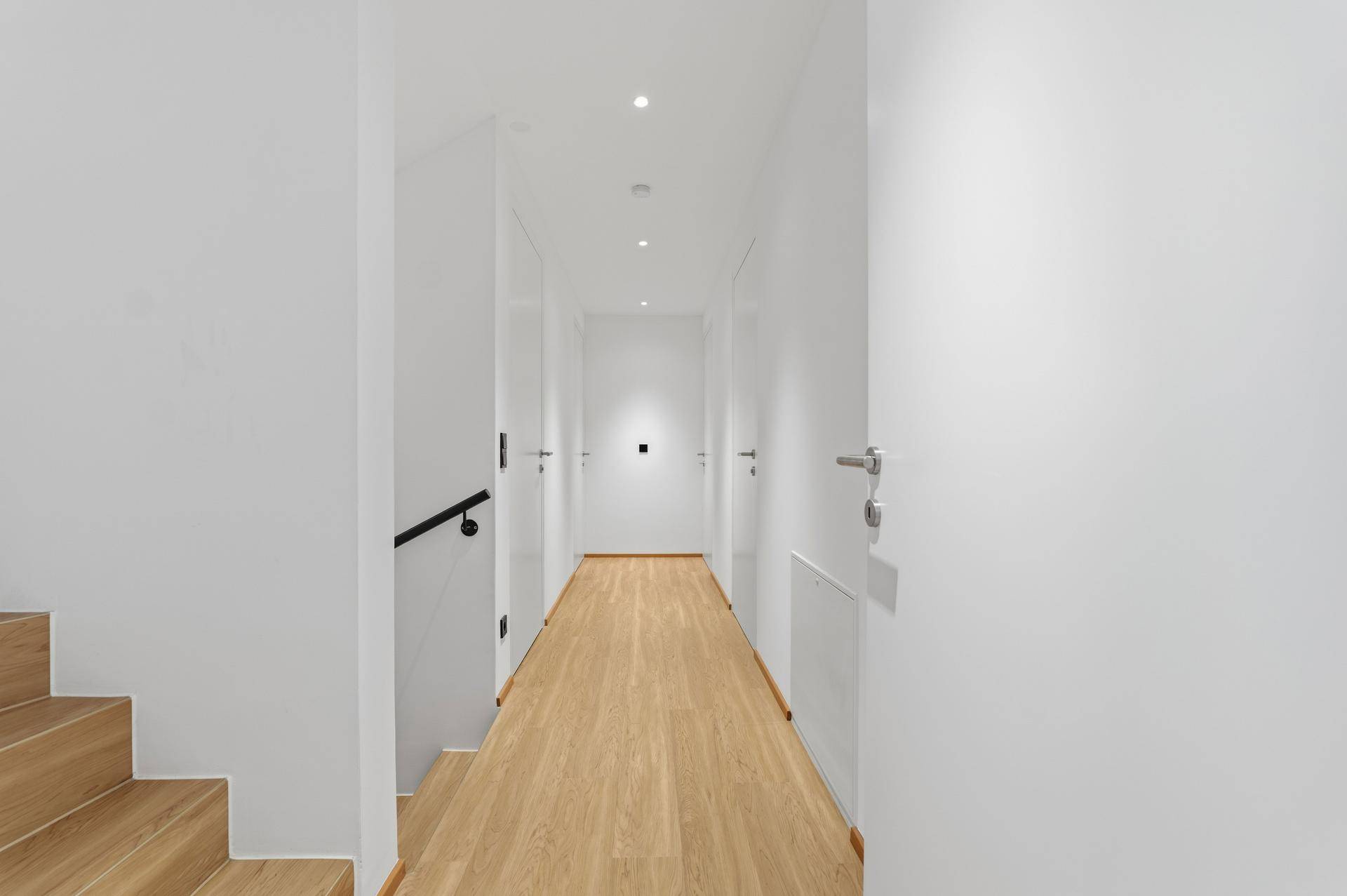 Heller Flur mit Holztreppe, weißen Wänden und integrierter Deckenbeleuchtung.