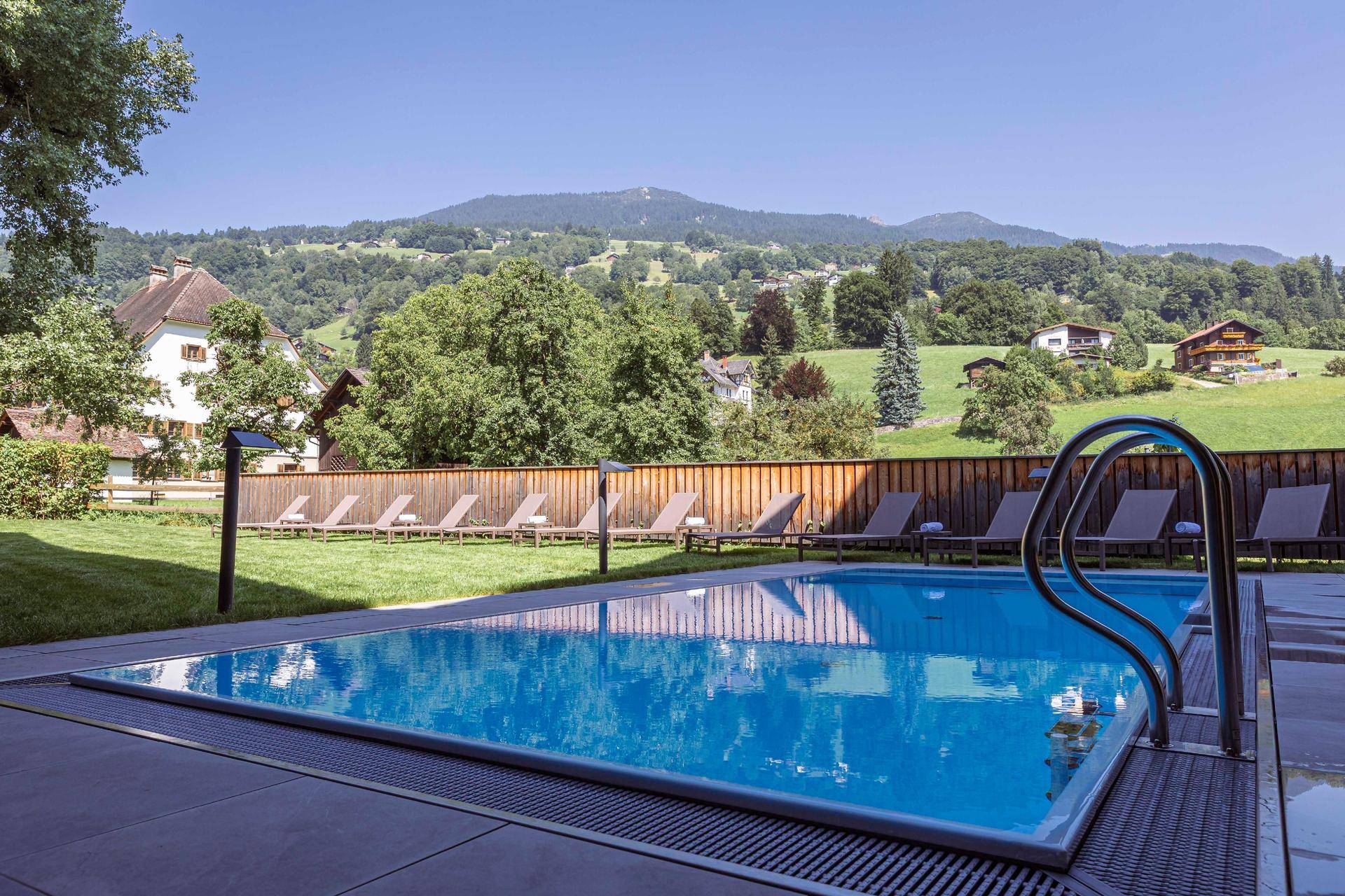 Außenpool mit Liegestühlen und einem weiten Blick auf die umliegende grüne Landschaft und Berge.