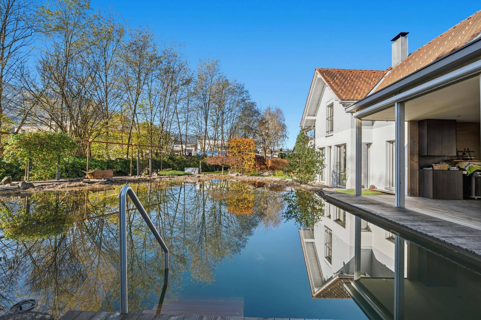 Hausansicht mit großem Naturpool und Holzdeck, umgeben von herbstlicher Vegetation unter blauem Himmel.