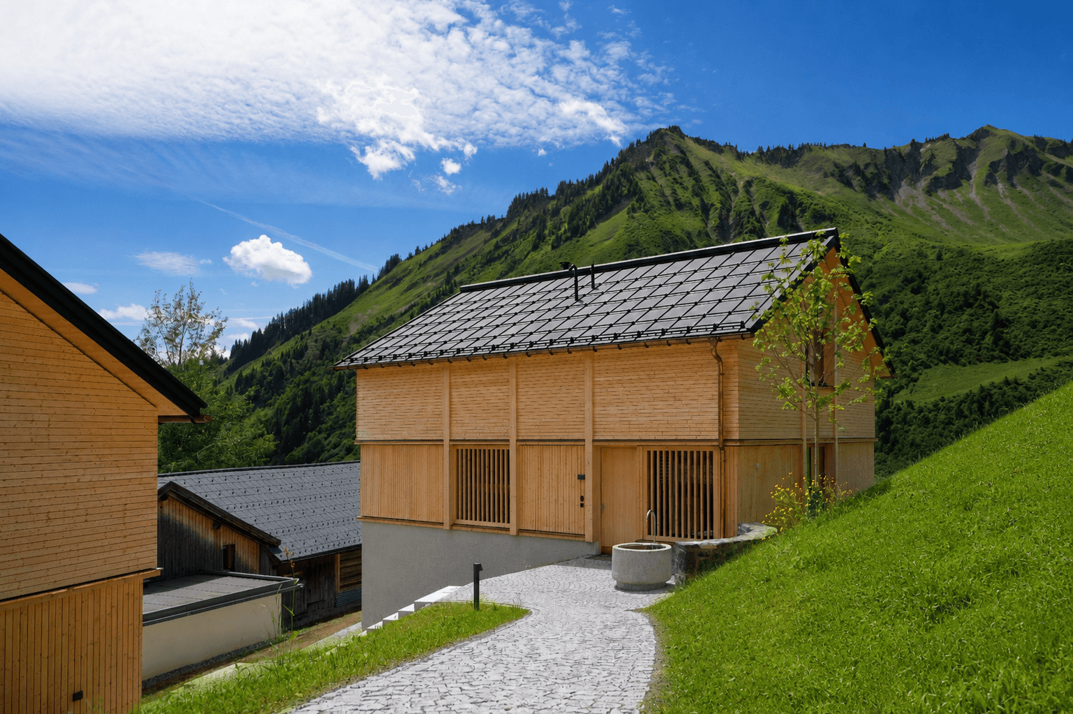 Neubau-Chalet mit Holzfassade und gepflastertem Weg in den Bergen unter strahlend blauem Himmel.