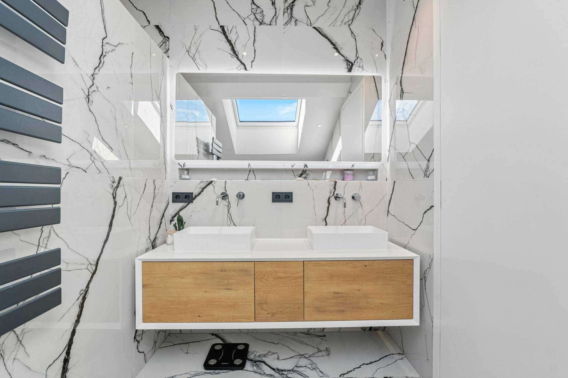 Modernes Badezimmer mit Marmoroptik-Fliesen, Doppelwaschbecken und beleuchtetem Spiegel unter einem Dachfenster.