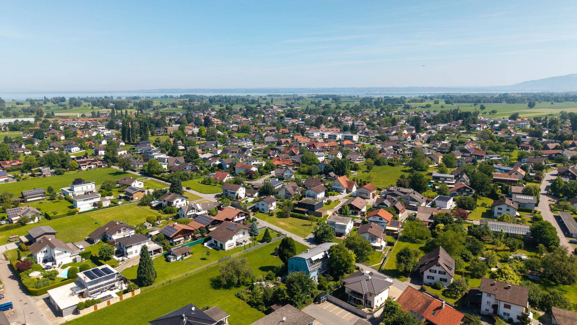 Luftaufnahme einer Wohnsiedlung mit vielen Häusern, grünen Feldern und einem See im Hintergrund.