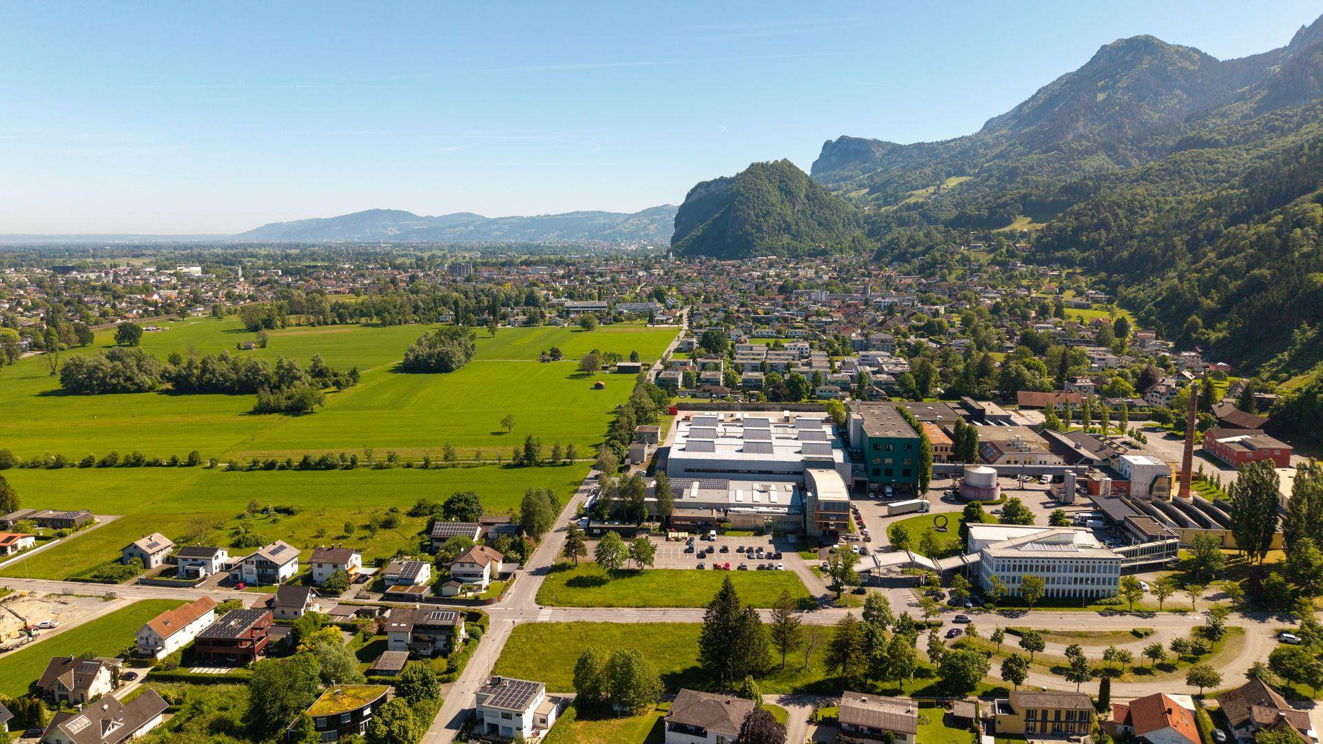 Weitwinkel-Luftaufnahme der Siedlung mit angrenzenden Feldern und einer beeindruckenden Bergkette im Hintergrund.