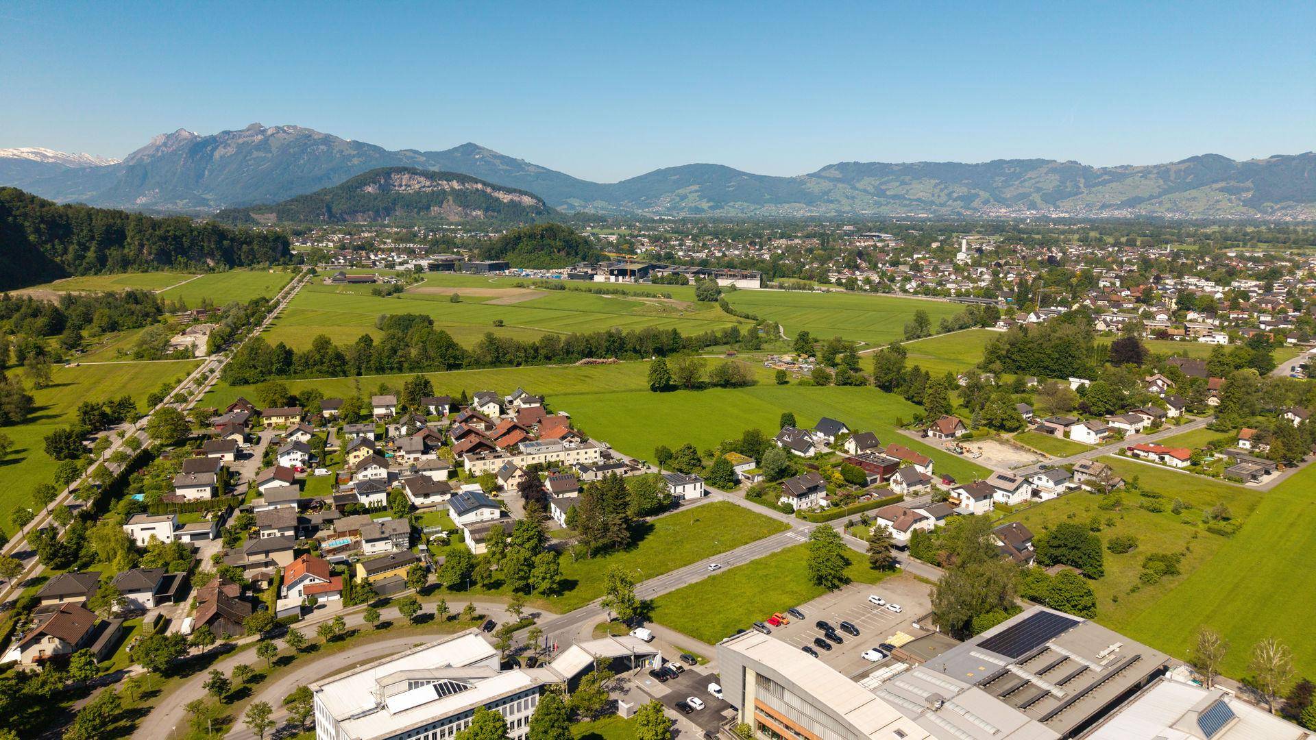 Weitwinkel-Luftaufnahme einer Gemeinde, umgeben von grünen Feldern und fernen Bergen.