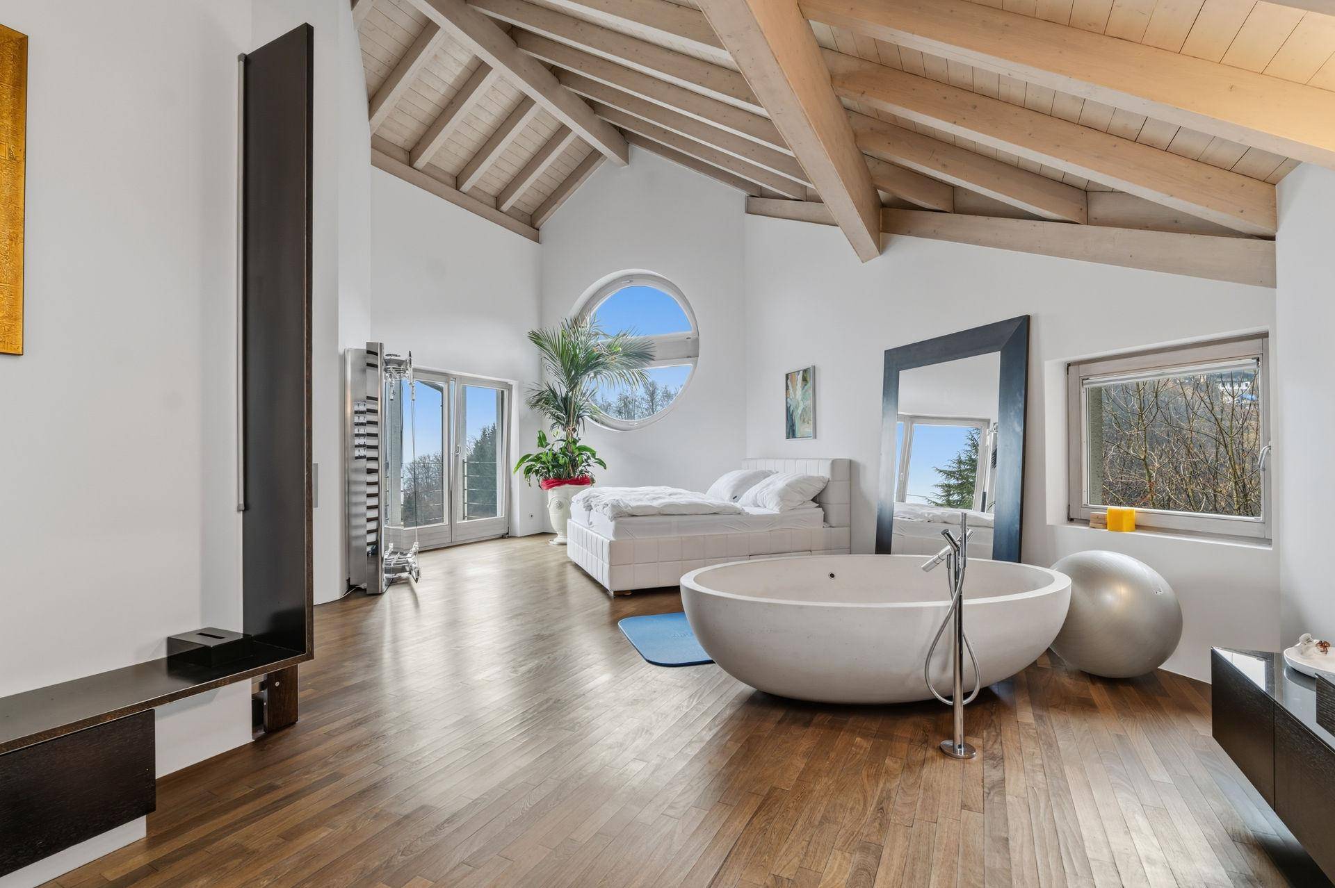 Helles Schlafzimmer mit freistehender Badewanne, Holzbalken und einem runden Fenster mit Weitblick.