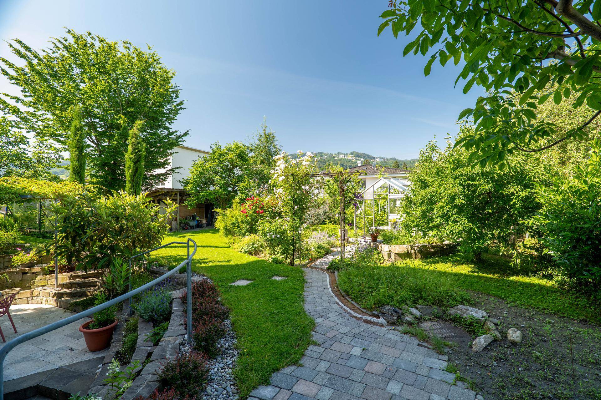 Weitläufiger, sonniger Garten mit Rasenflächen, Bäumen und einem gepflasterten Weg.