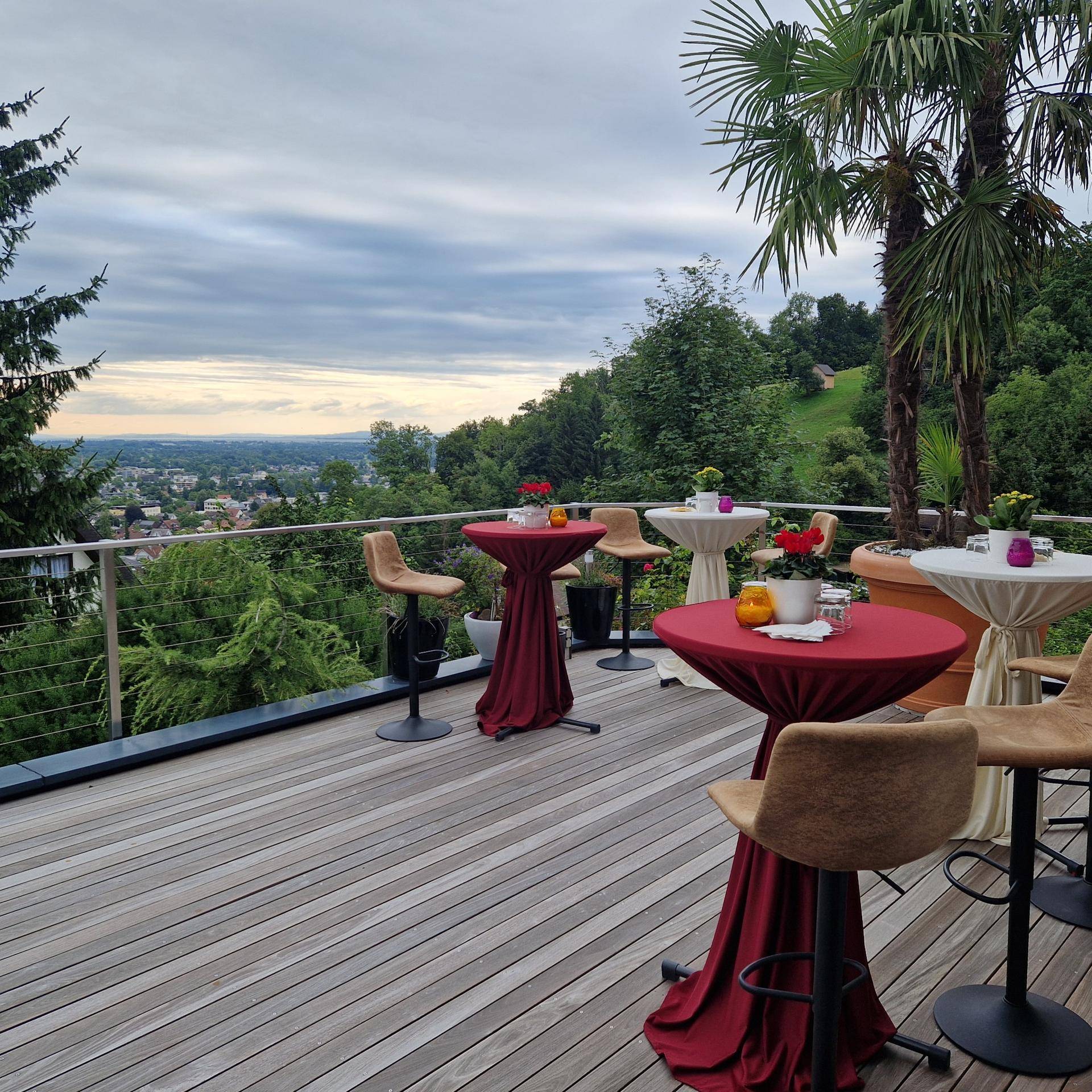 Große Holzterrasse mit Stehtischen und Stühlen, umgeben von Palmen und mit weitem Ausblick.