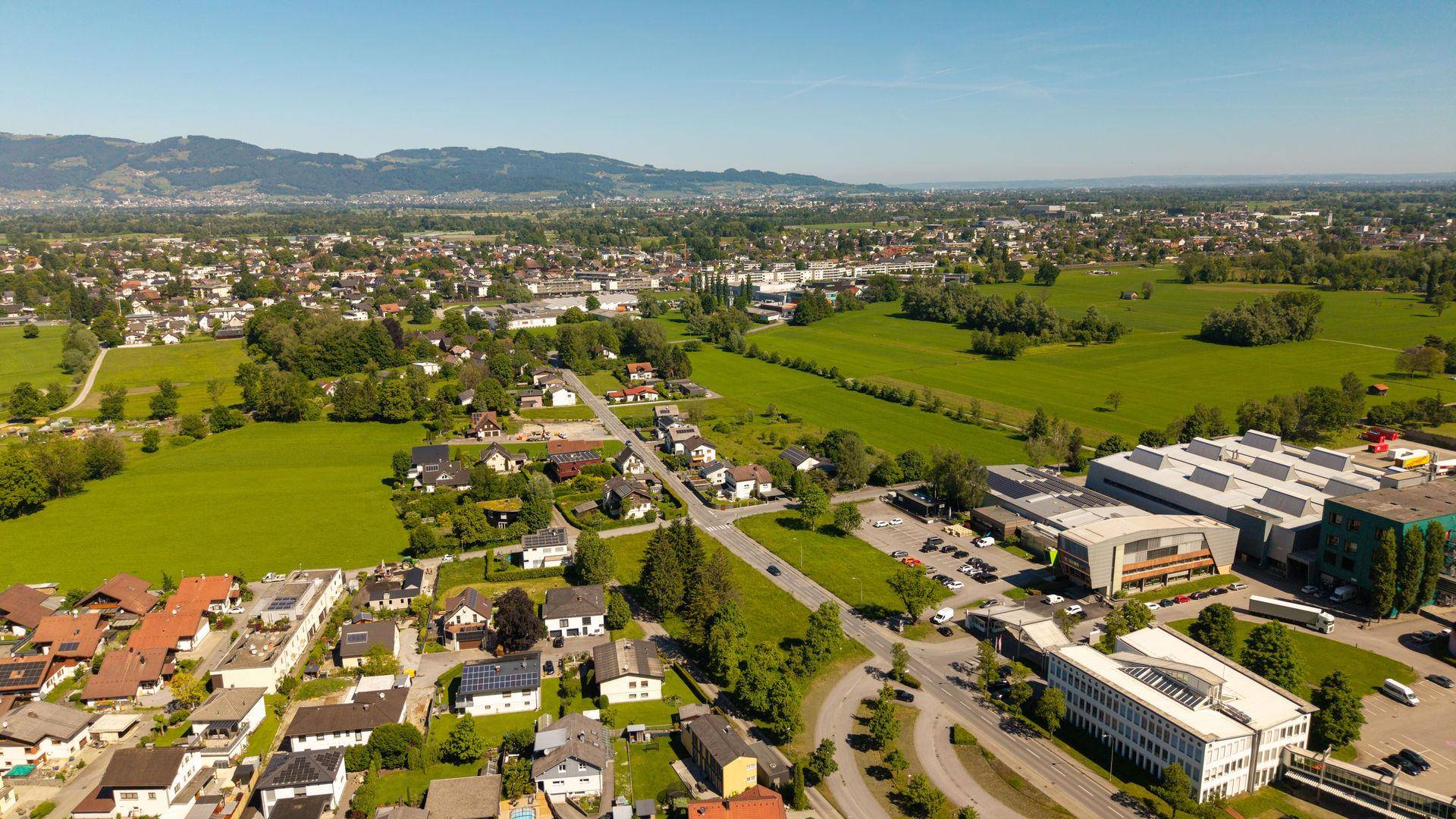 Panoramische Luftaufnahme der Umgebung mit Wohngebieten, Gewerbeflächen und grünen Feldern vor Bergkulisse.