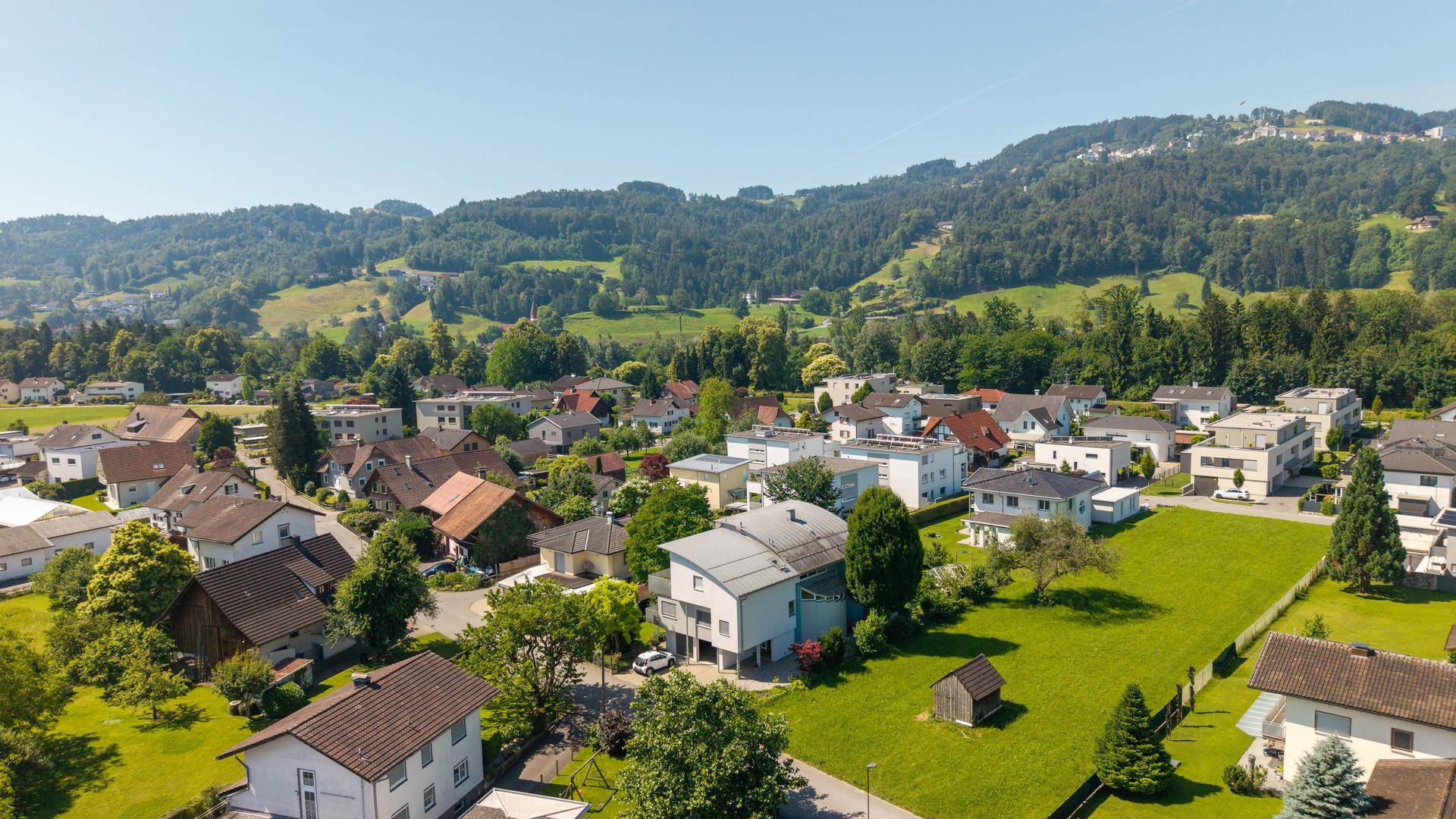 Luftaufnahme einer Wohnsiedlung mit Häusern und bewaldeten Hügeln im Hintergrund unter blauem Himmel.