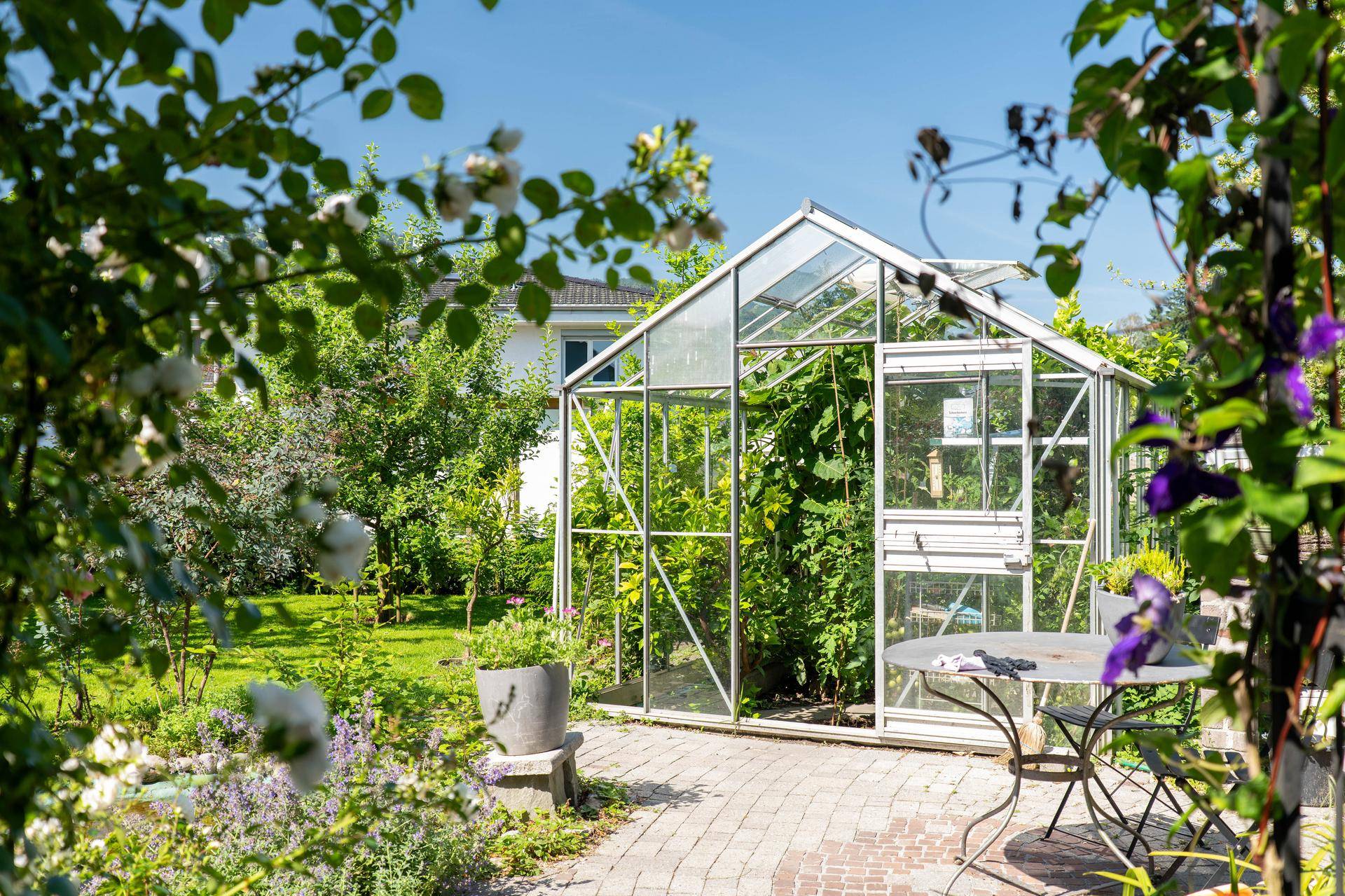 Gepflegter Garten mit Gewächshaus und kleinem Sitzbereich, umgeben von blühenden Pflanzen.