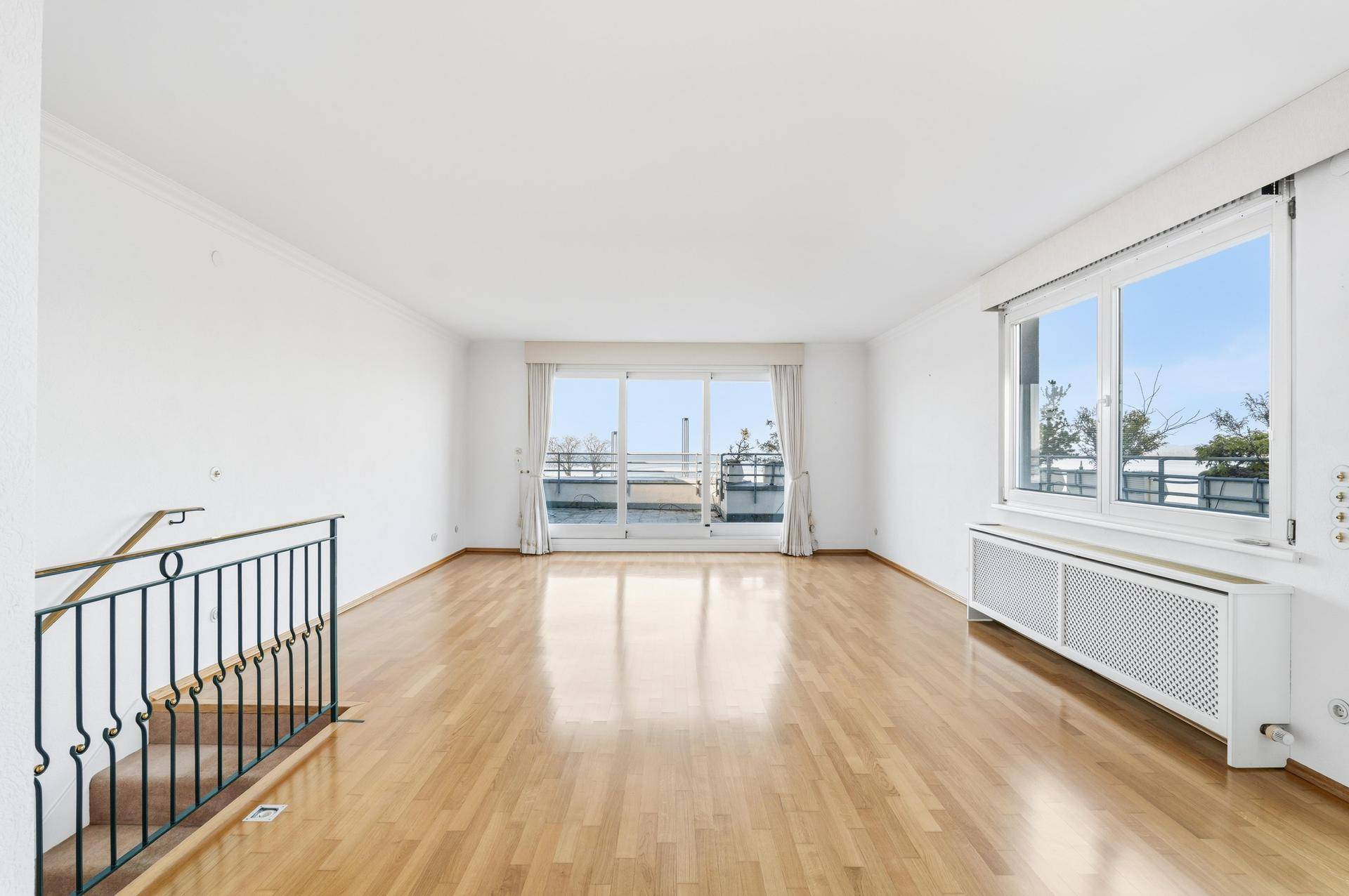 Helles Wohnzimmer mit glänzendem Parkettboden, großen Fenstern und Zugang zu einem Balkon mit Seeblick.