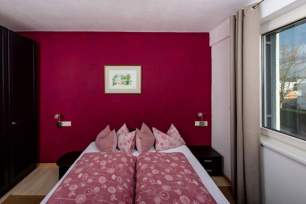 Schlafzimmer mit auffälliger roter Akzentwand, Doppelbett und Fenster für Tageslicht.