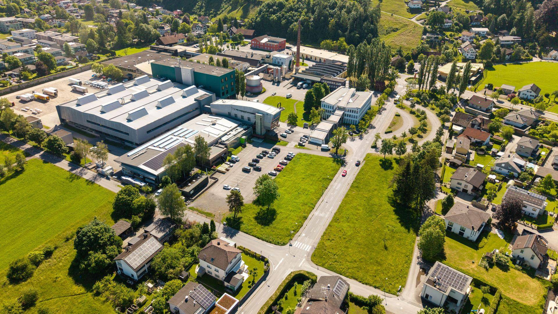 Luftaufnahme einer Ortschaft mit Wohn- und Industriegebäuden, umgeben von Natur und Bergen.