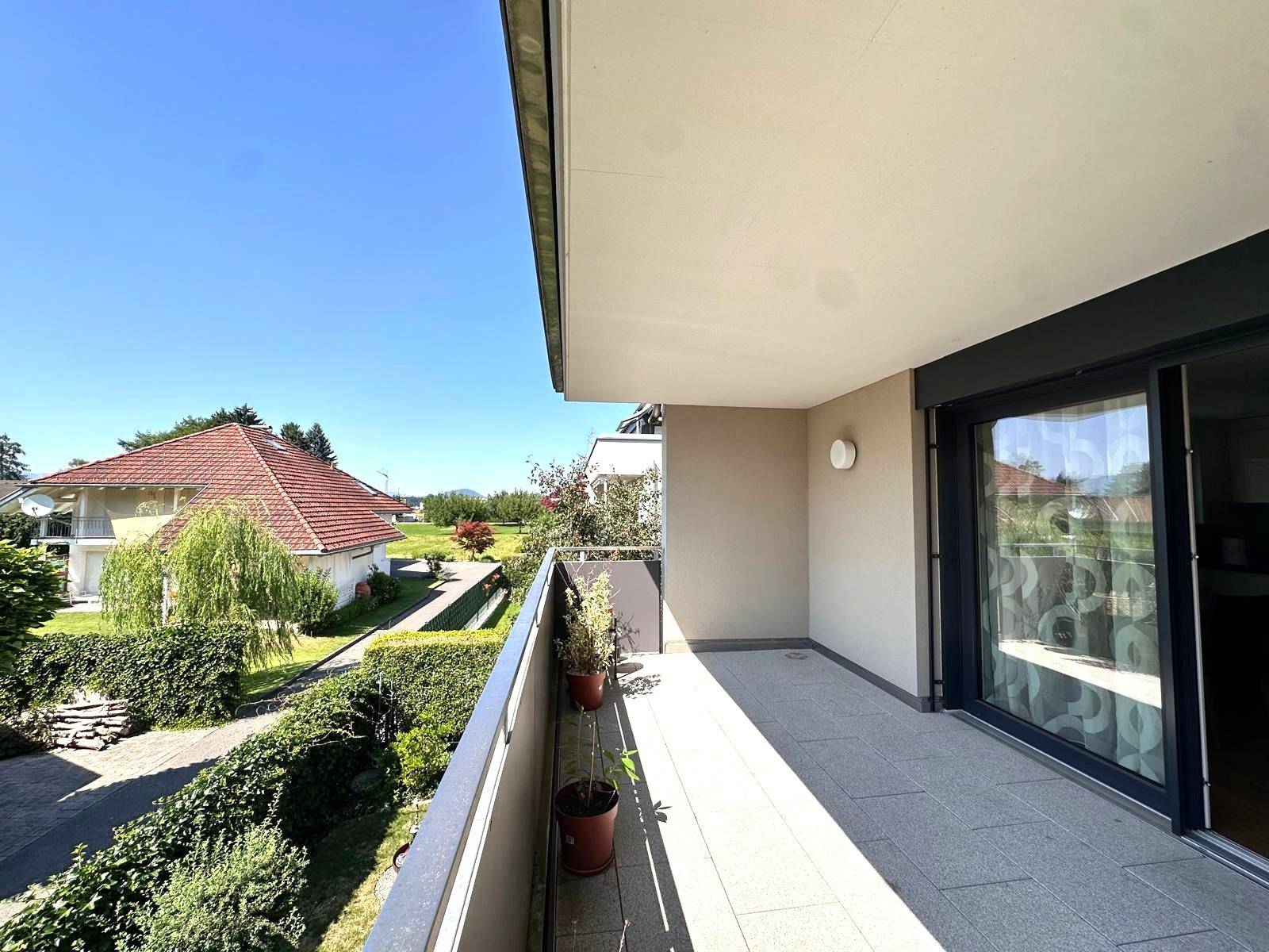 Heller Balkon mit Fliesenboden und Blick auf die grüne Umgebung und Nachbarhäuser unter blauem Himmel.