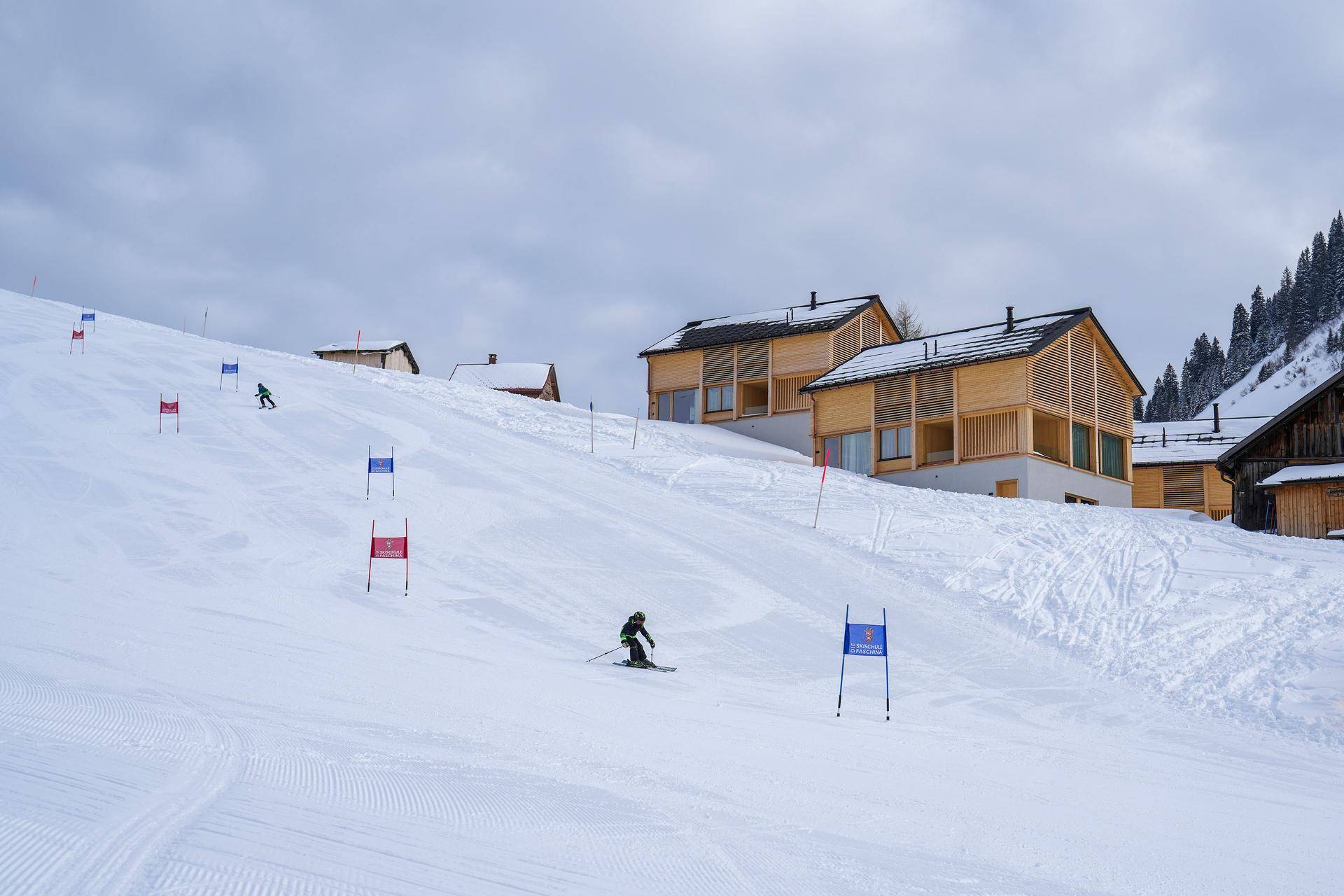 Moderne Holzhäuser an einem verschneiten Berghang mit Skifahrern auf der Piste.