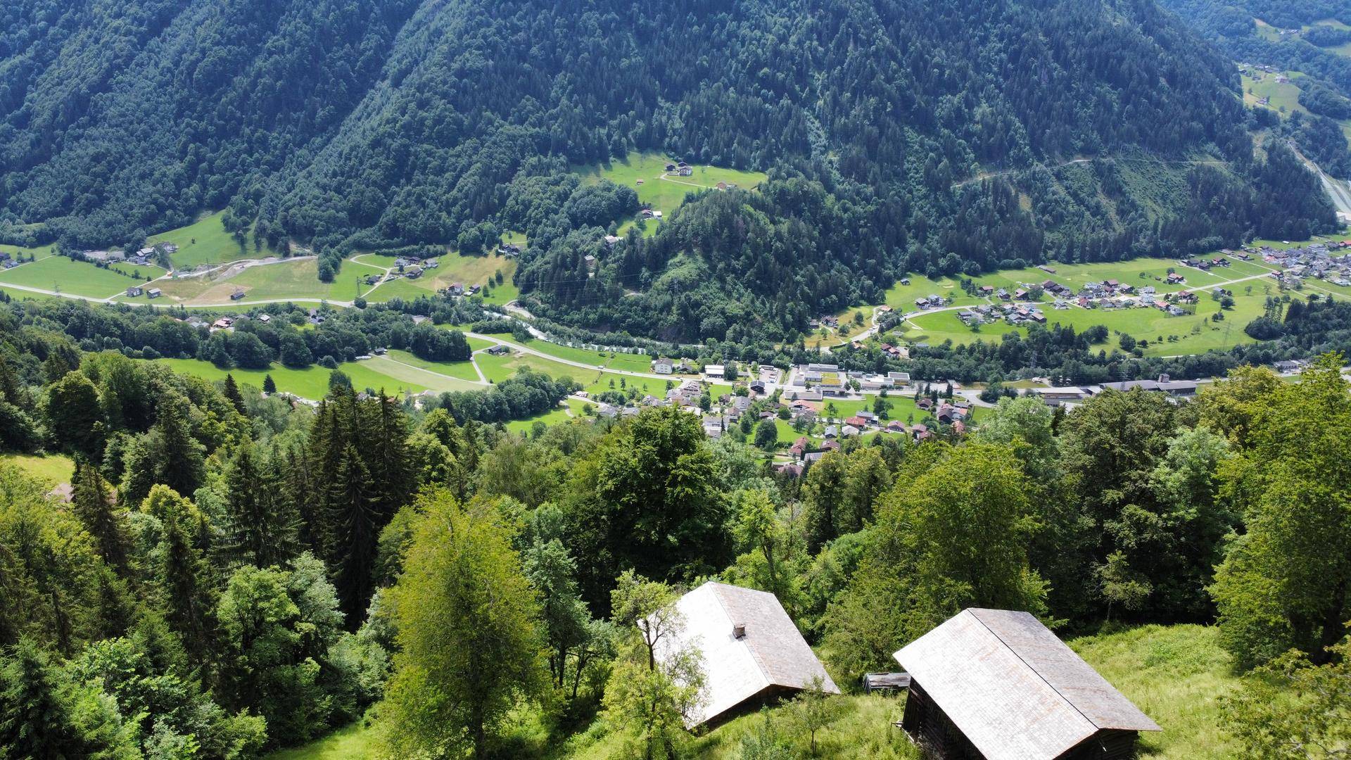 Weitläufiger Blick über ein grünes Tal mit bewaldeten Bergen und einem Dorf in der Ferne.