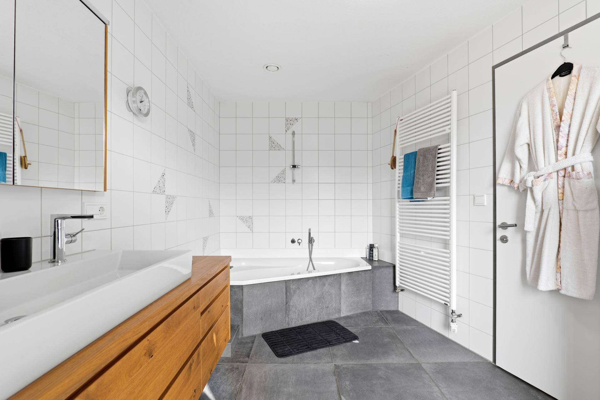 Modernes Badezimmer mit weißen Wandfliesen, grauen Bodenfliesen und einer Eckbadewanne.
