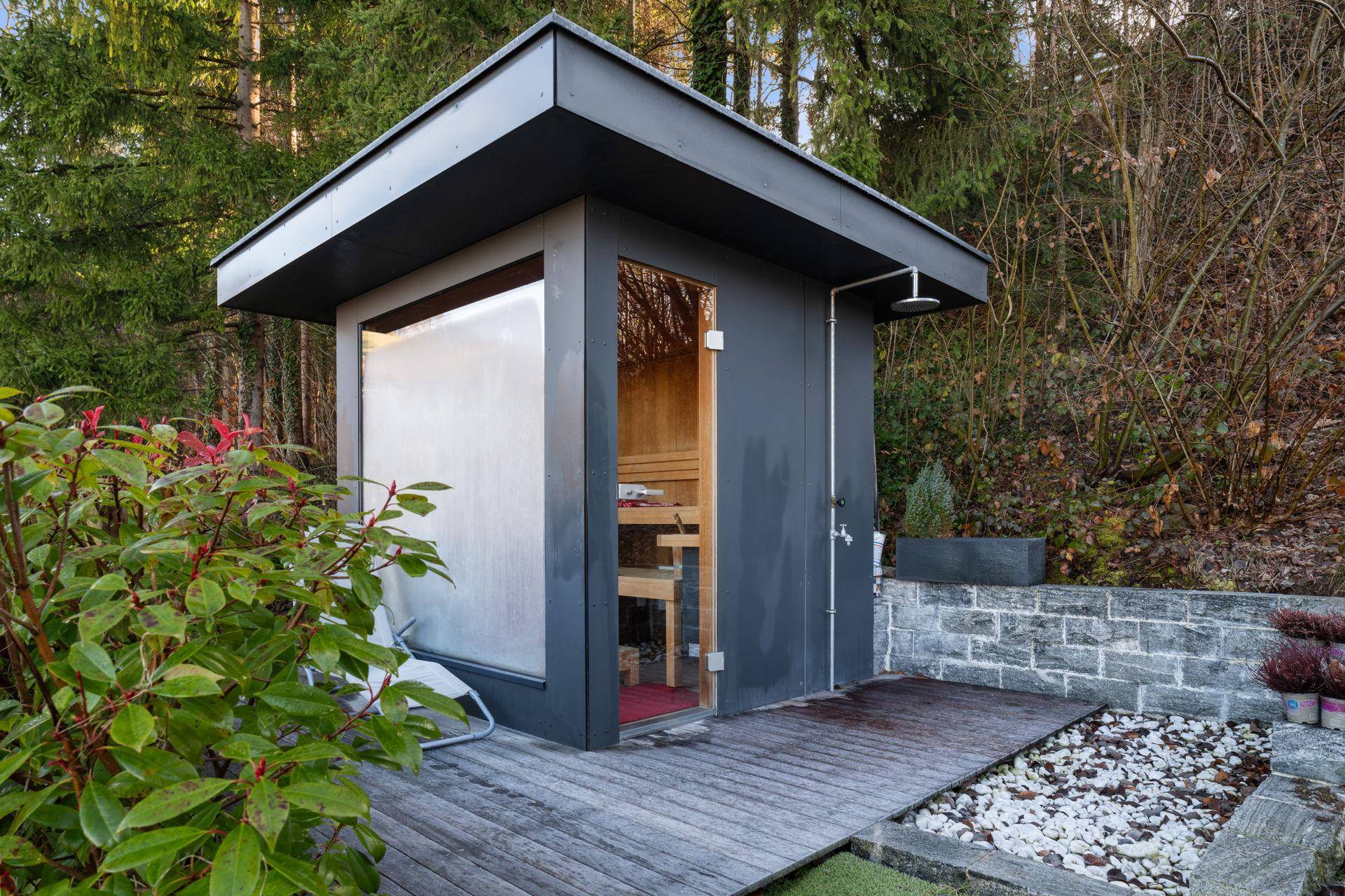 Moderne Außensauna im Gartenbereich mit Holzdeck und Außendusche, umgeben von Bäumen.