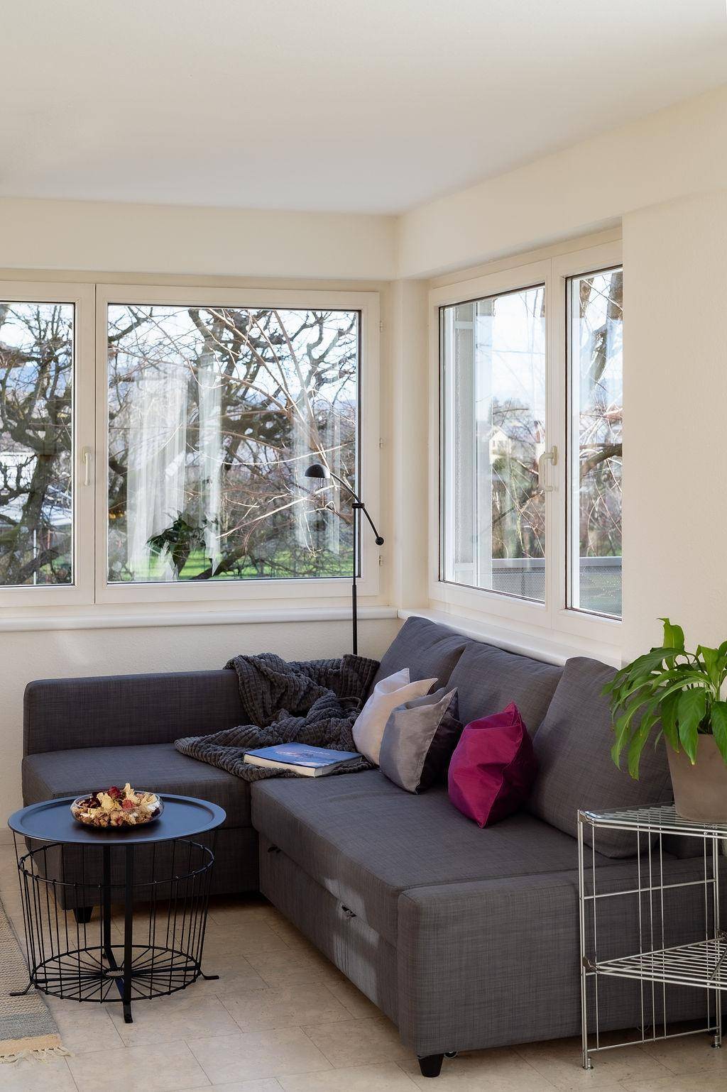 Helles Wohnzimmer mit grauem Ecksofa, modernem Couchtisch und großen Fenstern mit Baumblick.