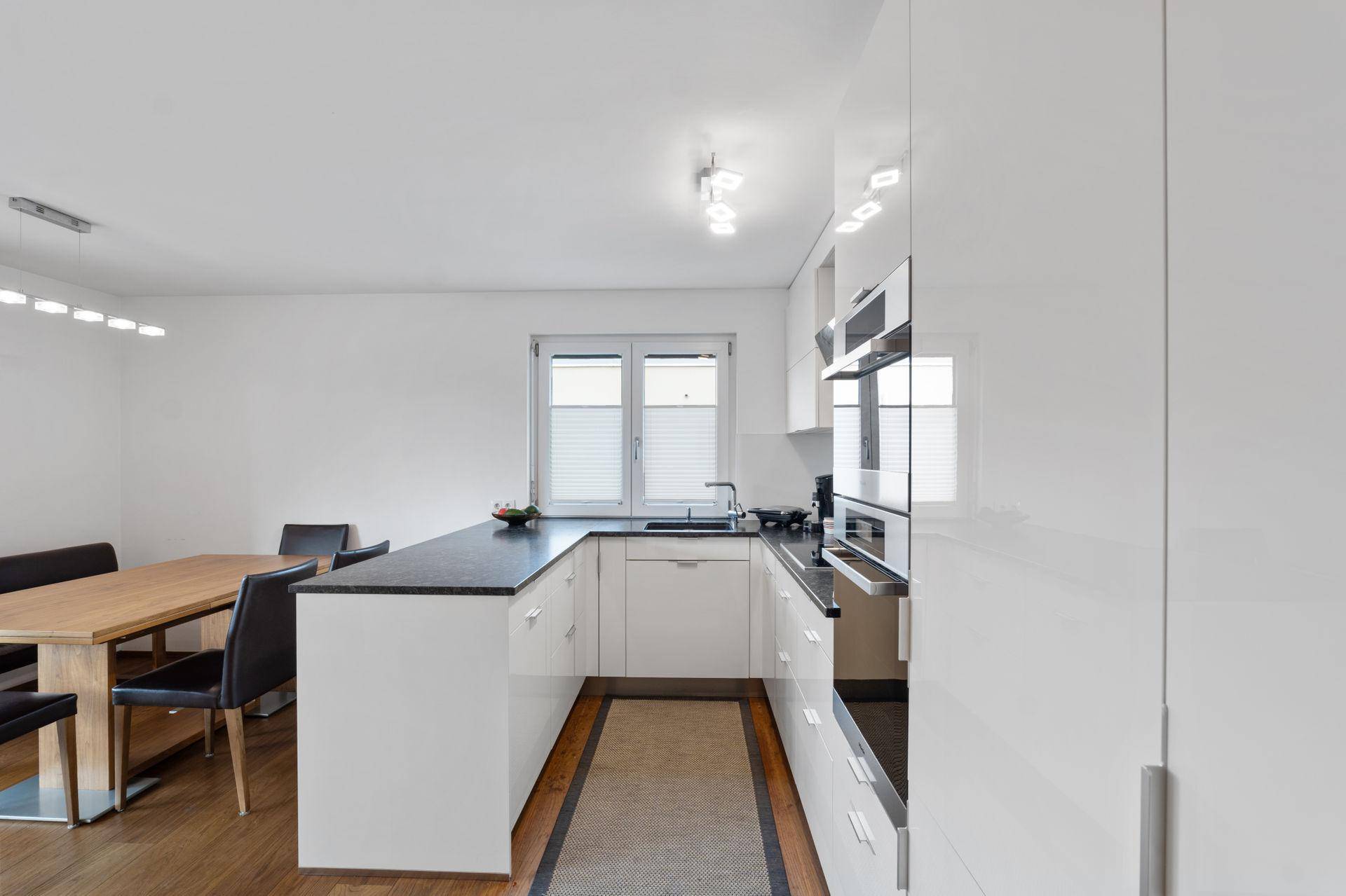 Moderne Küche mit weißen Hochglanzfronten, integriertem Essbereich und Fenster für natürliches Licht.