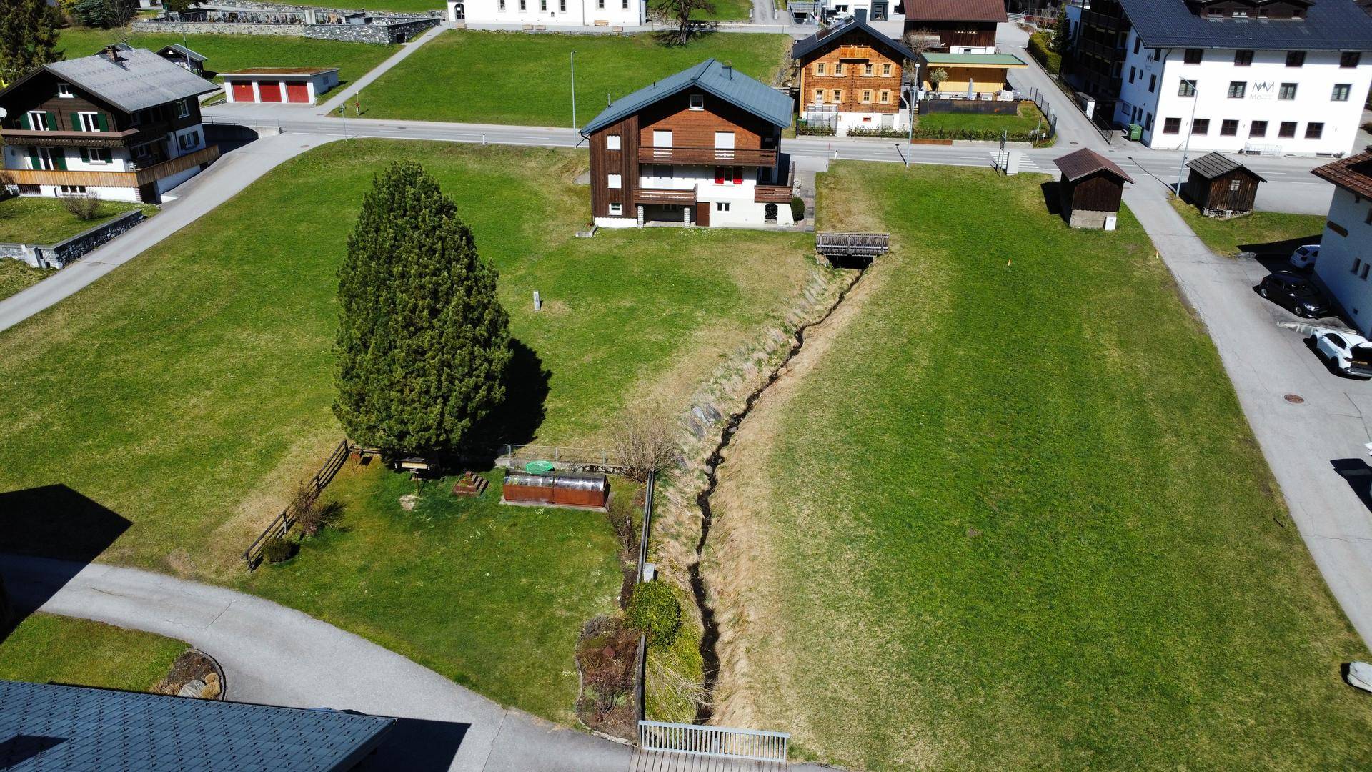 Luftaufnahme eines Einfamilienhauses mit Balkonen, umgeben von grünen Wiesen und einem Bachlauf.
