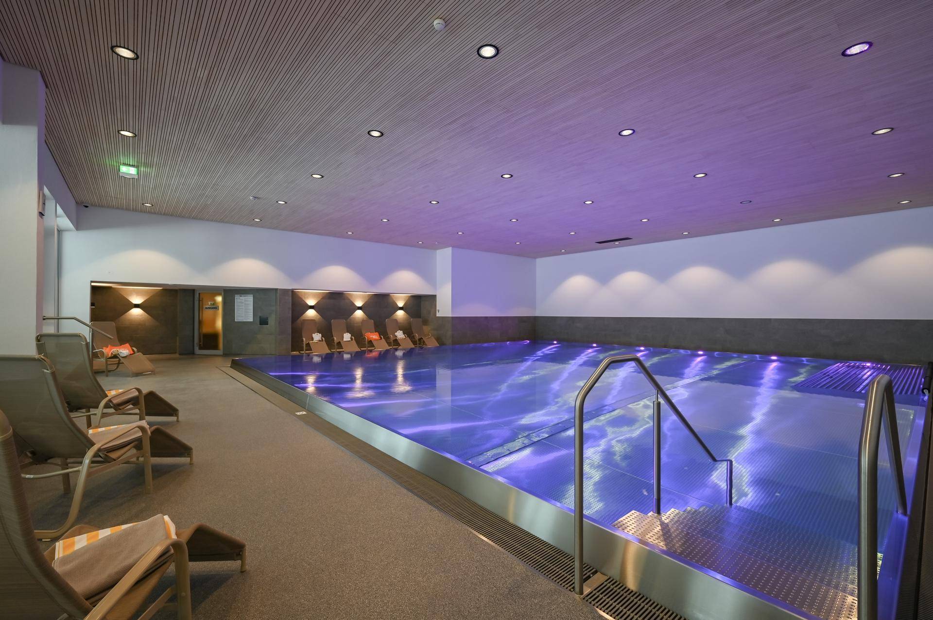 Modernes Hallenbad mit blauer Beleuchtung und bequemen Liegestühlen zum Entspannen.
