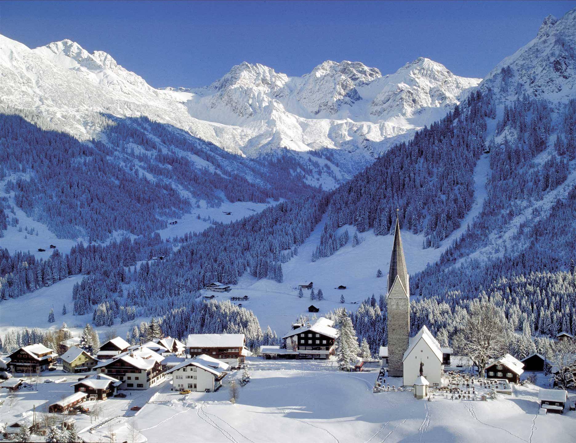 Malerisches, schneebedecktes Dorf mit Kirche, umgeben von majestätischen Bergen und Wäldern.