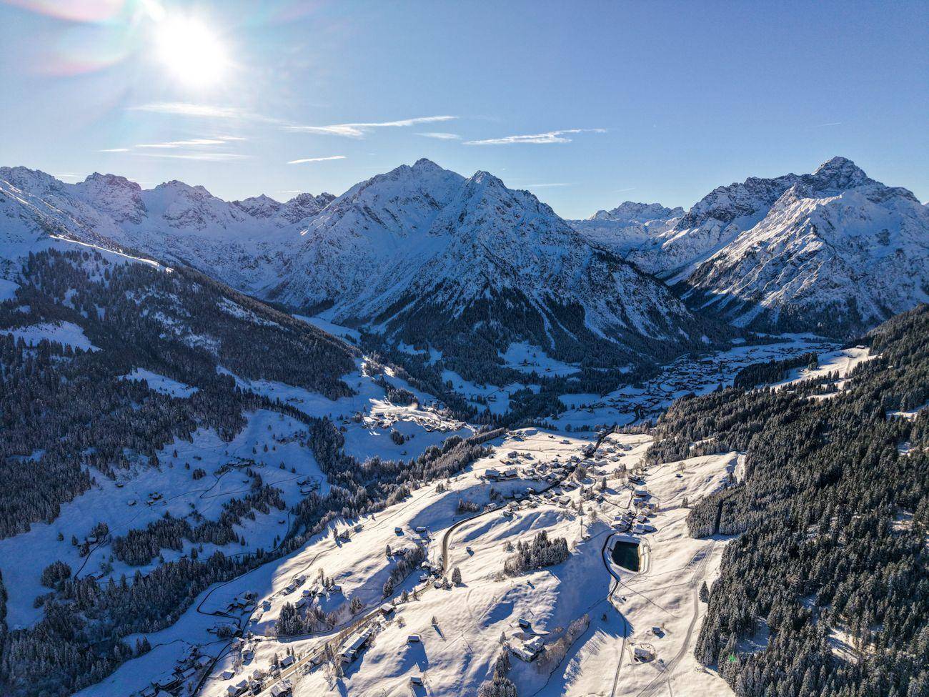 Atemberaubende Luftaufnahme eines schneebedeckten Tals mit einem Dorf und majestätischen Bergen unter strahlendem Sonnenschein.