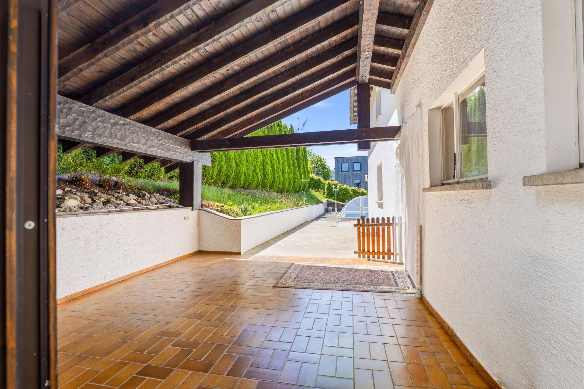 Überdachte Loggia mit Fliesenboden und Holzdach, die zum Gartenbereich führt.